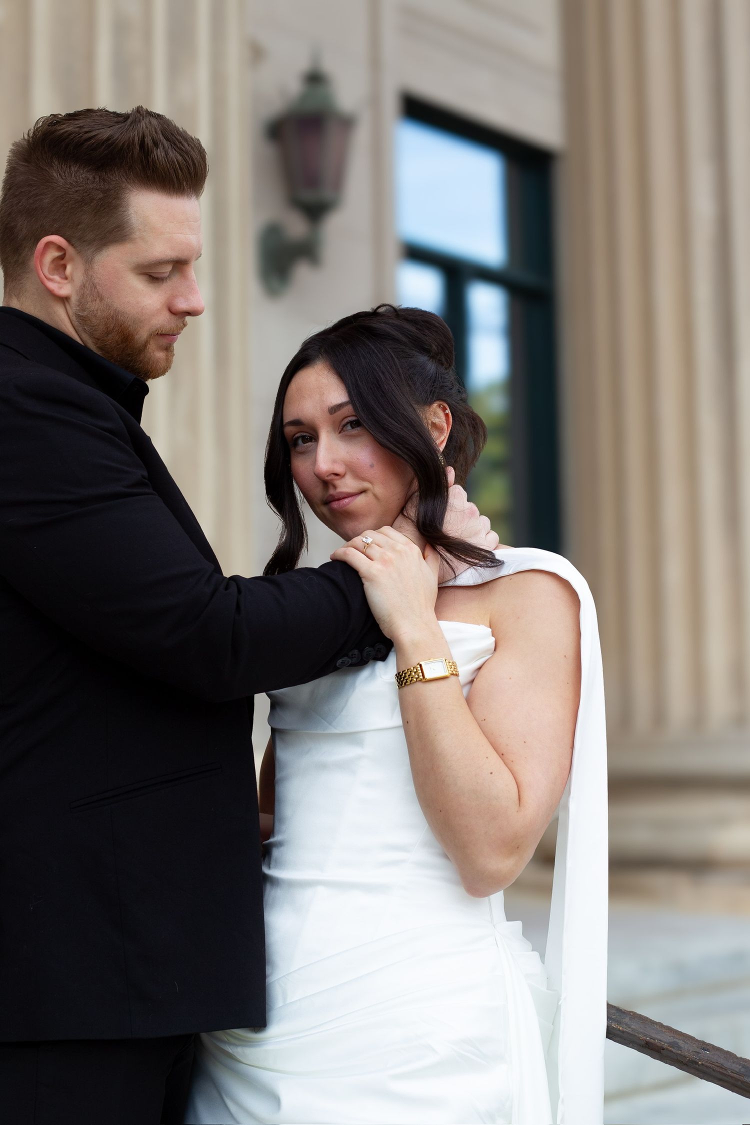 Groom in black suit tenderly touches bride's face as she holds his hand, standing before elegant columns.