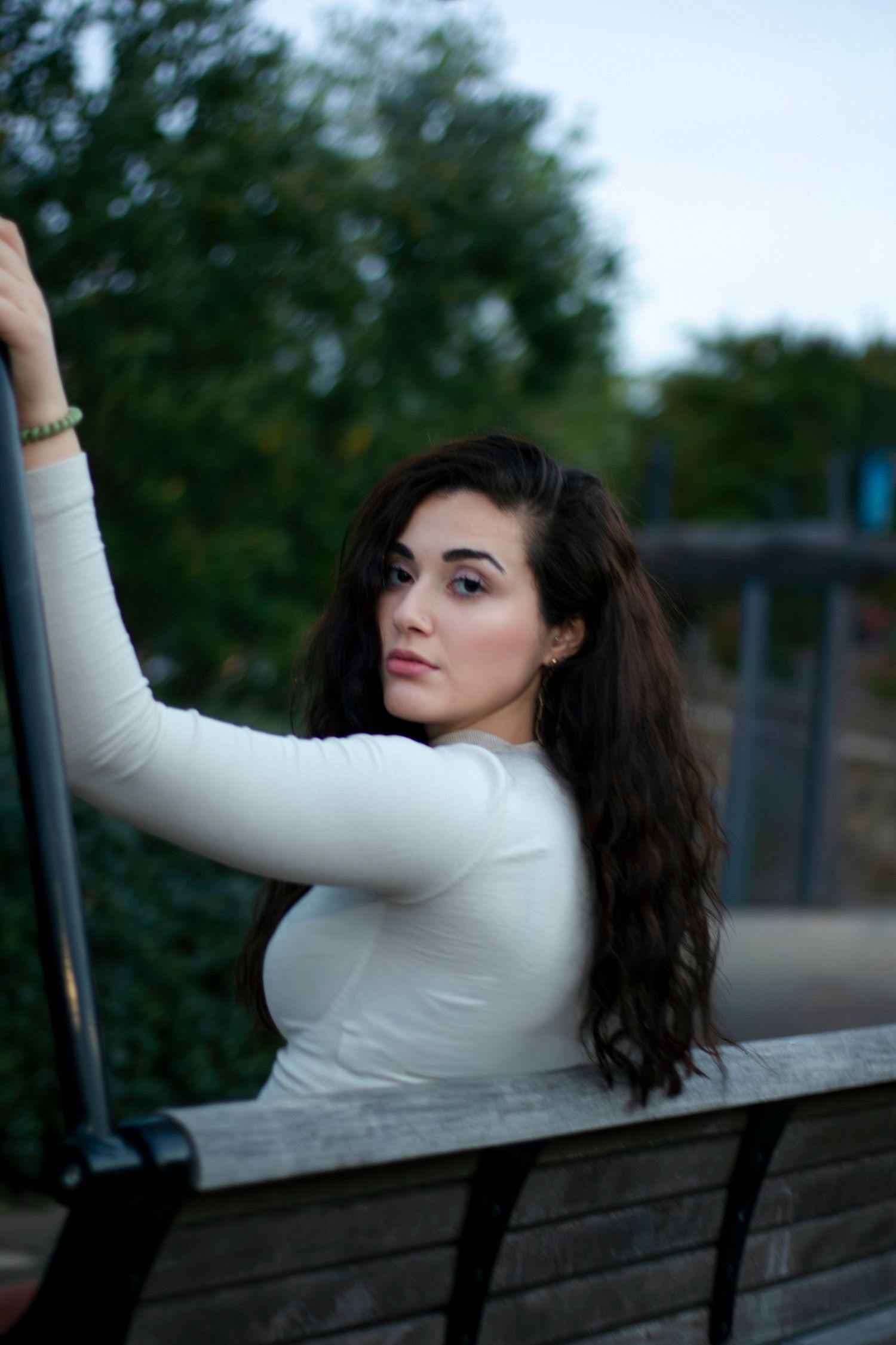 Young woman with long dark hair in white top poses on a bench outdoors with green trees in background.