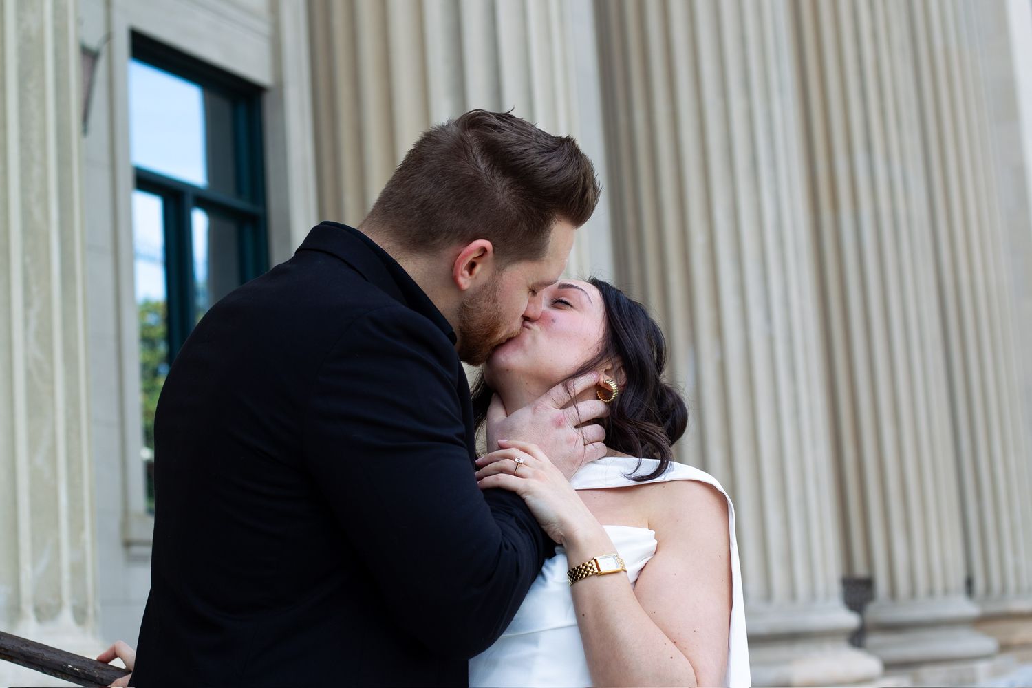 Couple sharing a romantic kiss in front of grand white columns, groom in black suit, bride in white dress with gold bracelet.
