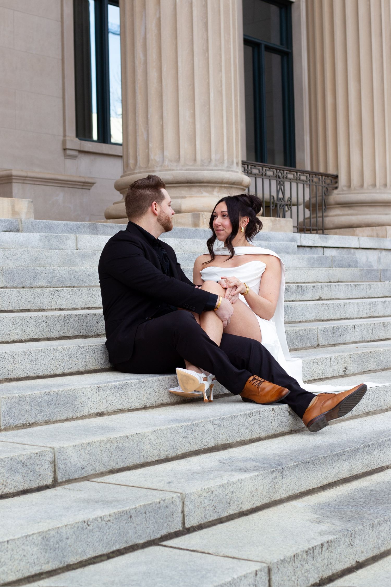 Bride and groom sitting on marble steps of a grand building, holding hands and smiling at each other.