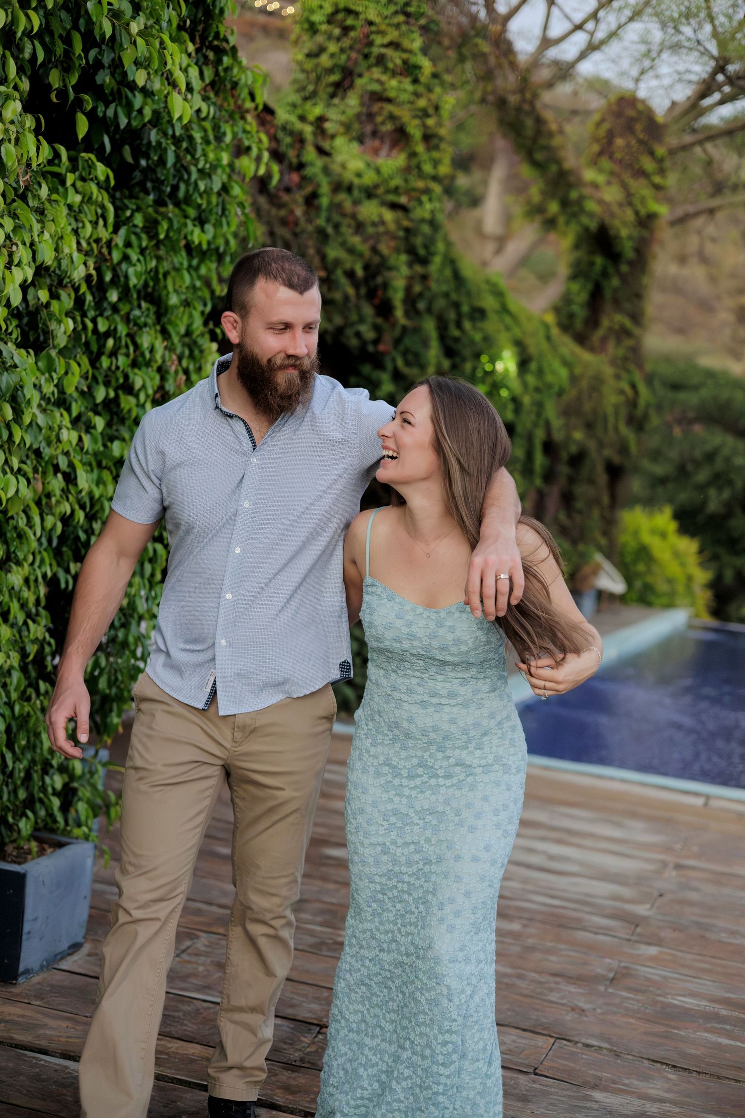 Happy couple walking together near a pool, woman in mint dress laughing, green ivy wall backdrop.