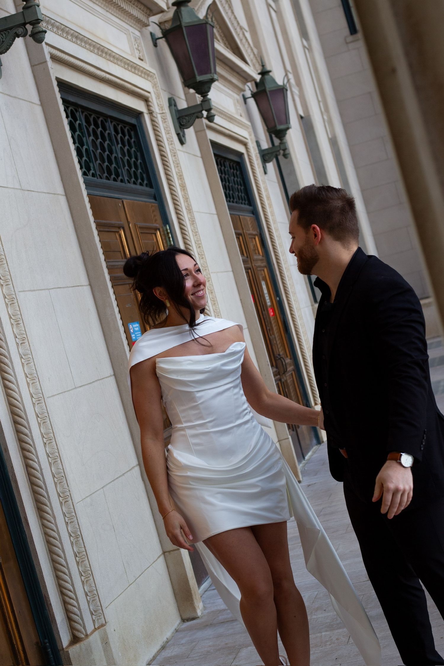 Couple in elegant attire sharing a romantic moment outside a classic building, woman in white mini dress, man in black suit.