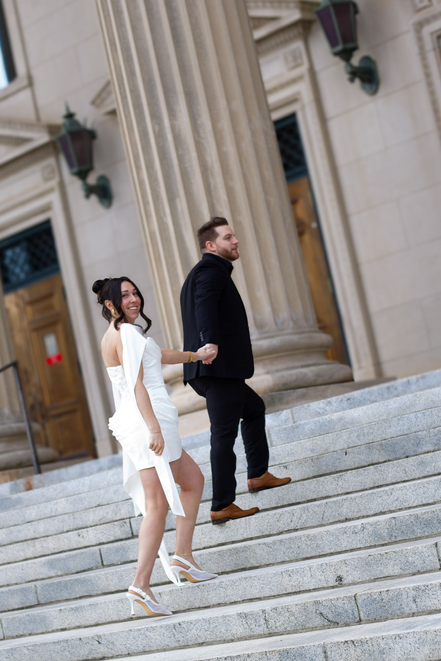 Bride in white dress and groom in black suit hold hands walking up stone steps outside a grand building with tall columns.