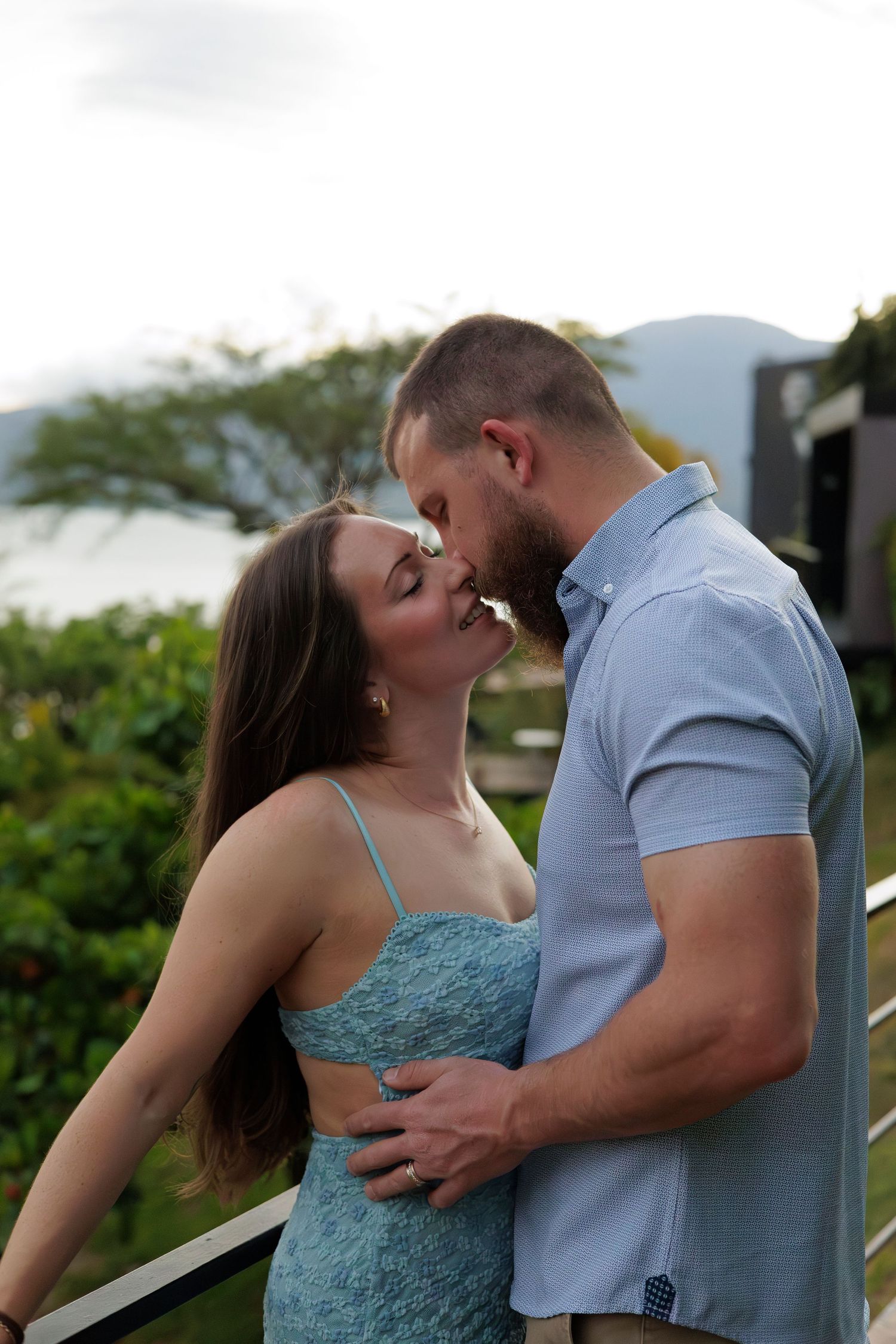A couple shares a romantic kiss on a balcony surrounded by lush tropical greenery and mountains.