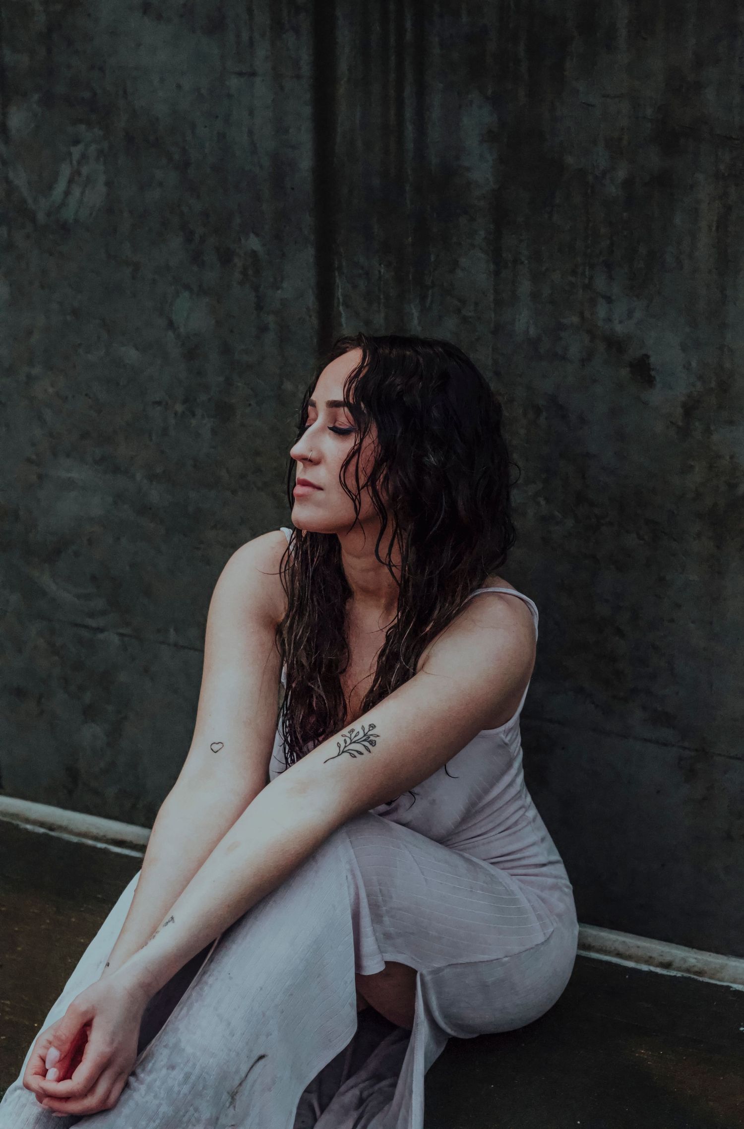 A woman with wet dark hair sits pensively in a white dress, showcasing a floral arm tattoo against a moody backdrop.