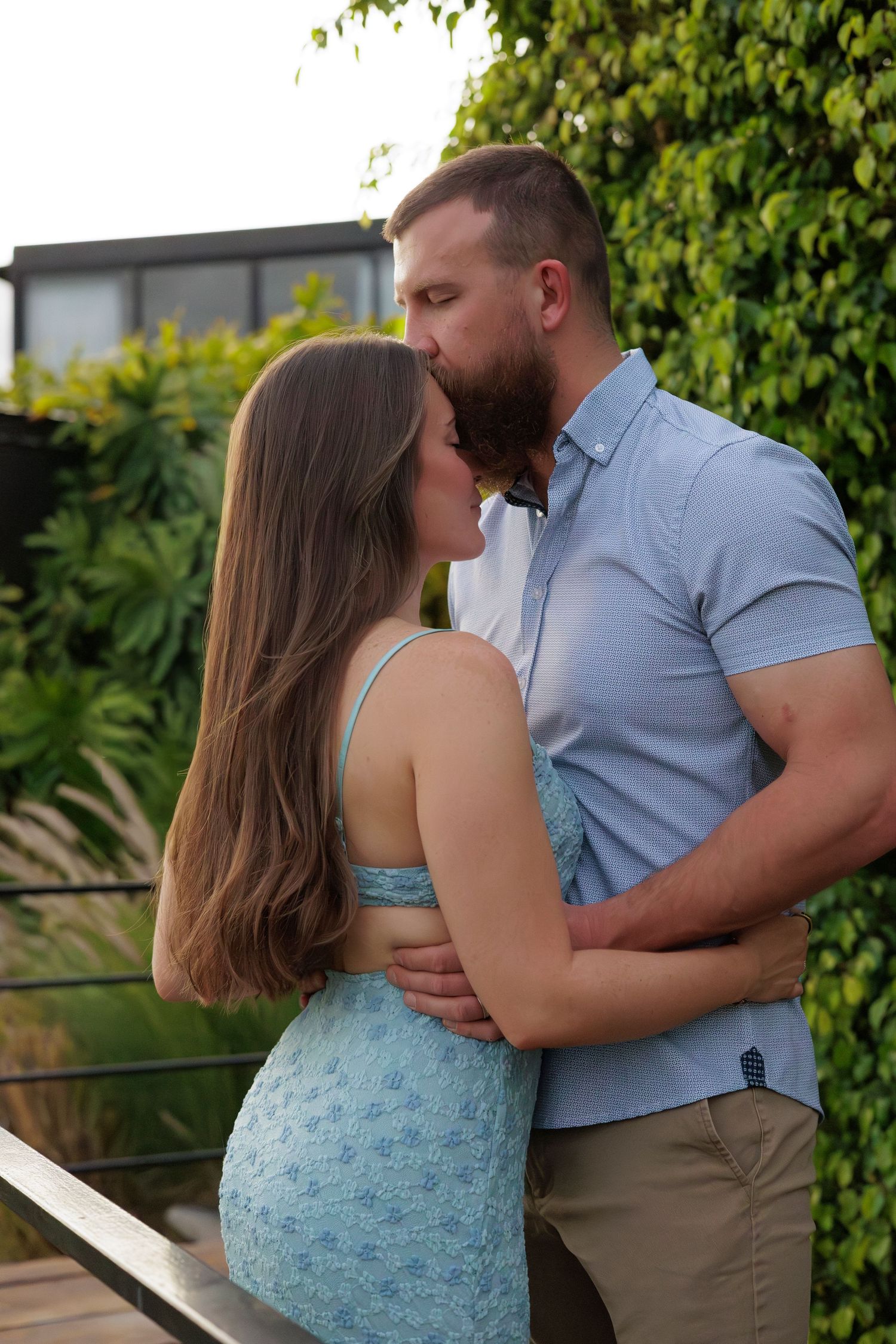 A couple sharing a romantic kiss on an outdoor balcony surrounded by lush green foliage.