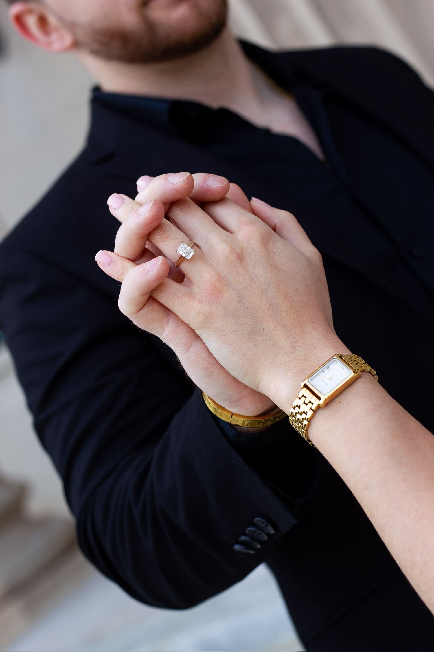 Couple holding hands showing engagement ring and gold watch, man in black suit jacket.