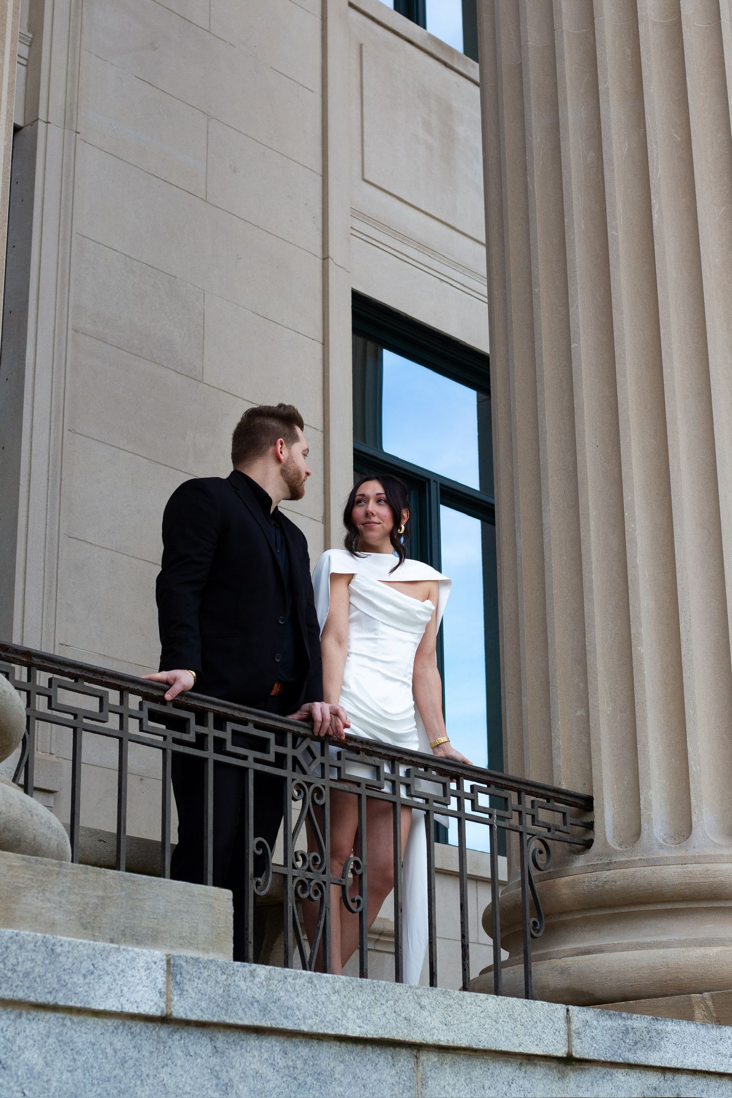 Elegant couple in formal attire standing on ornate building balcony, man in black suit, woman in white dress.