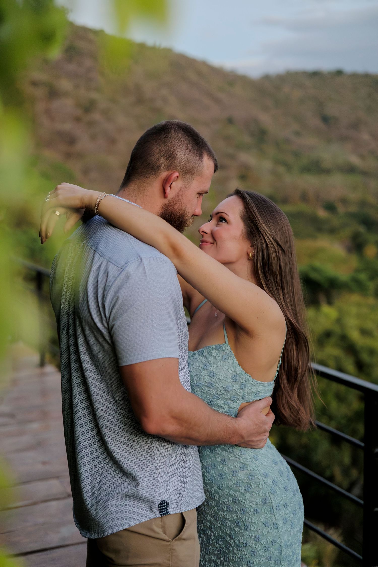 Couple embracing and gazing at each other on a scenic overlook with lush green hills in the background.