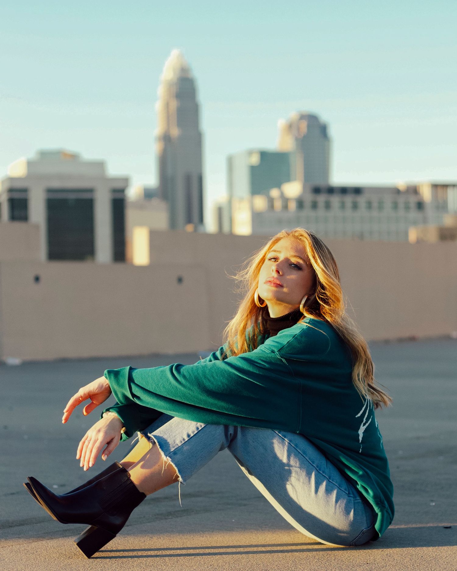 Young woman in green jacket and jeans sitting on rooftop with city skyline in background.