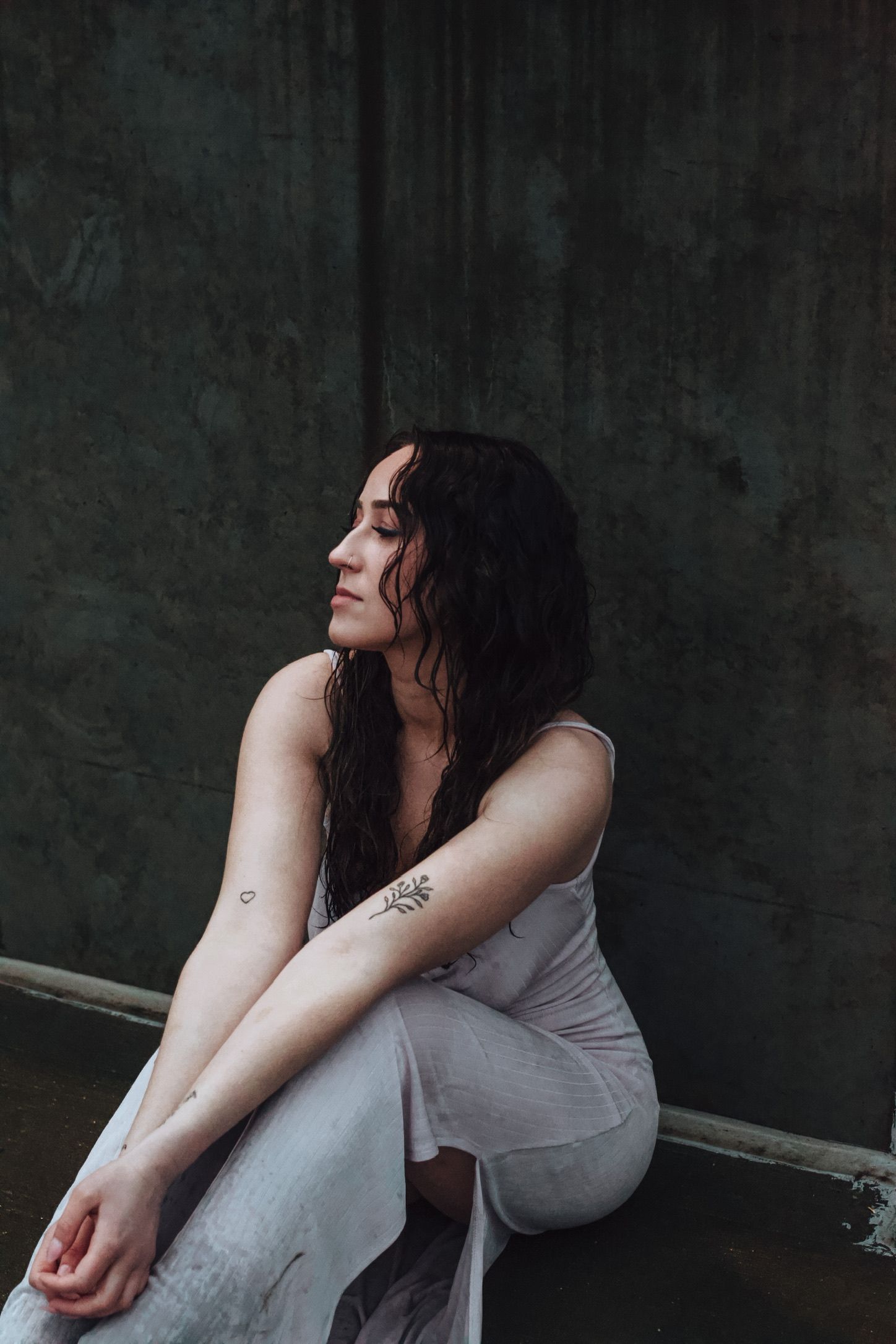Woman with wet dark hair and floral arm tattoo sits pensively in white dress against dark textured wall.