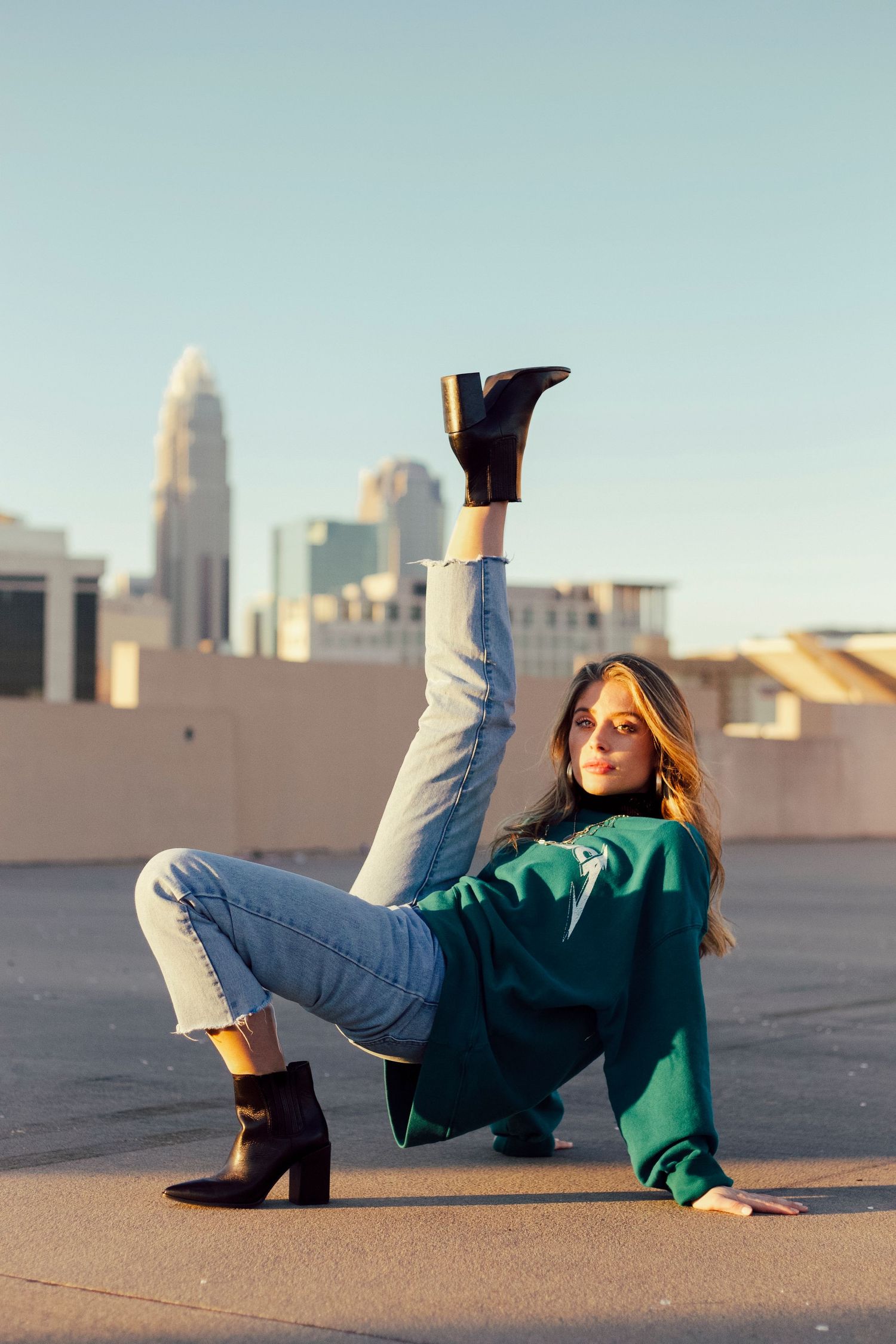 Woman in green sweatshirt and jeans poses on rooftop with leg raised, city skyline in background.