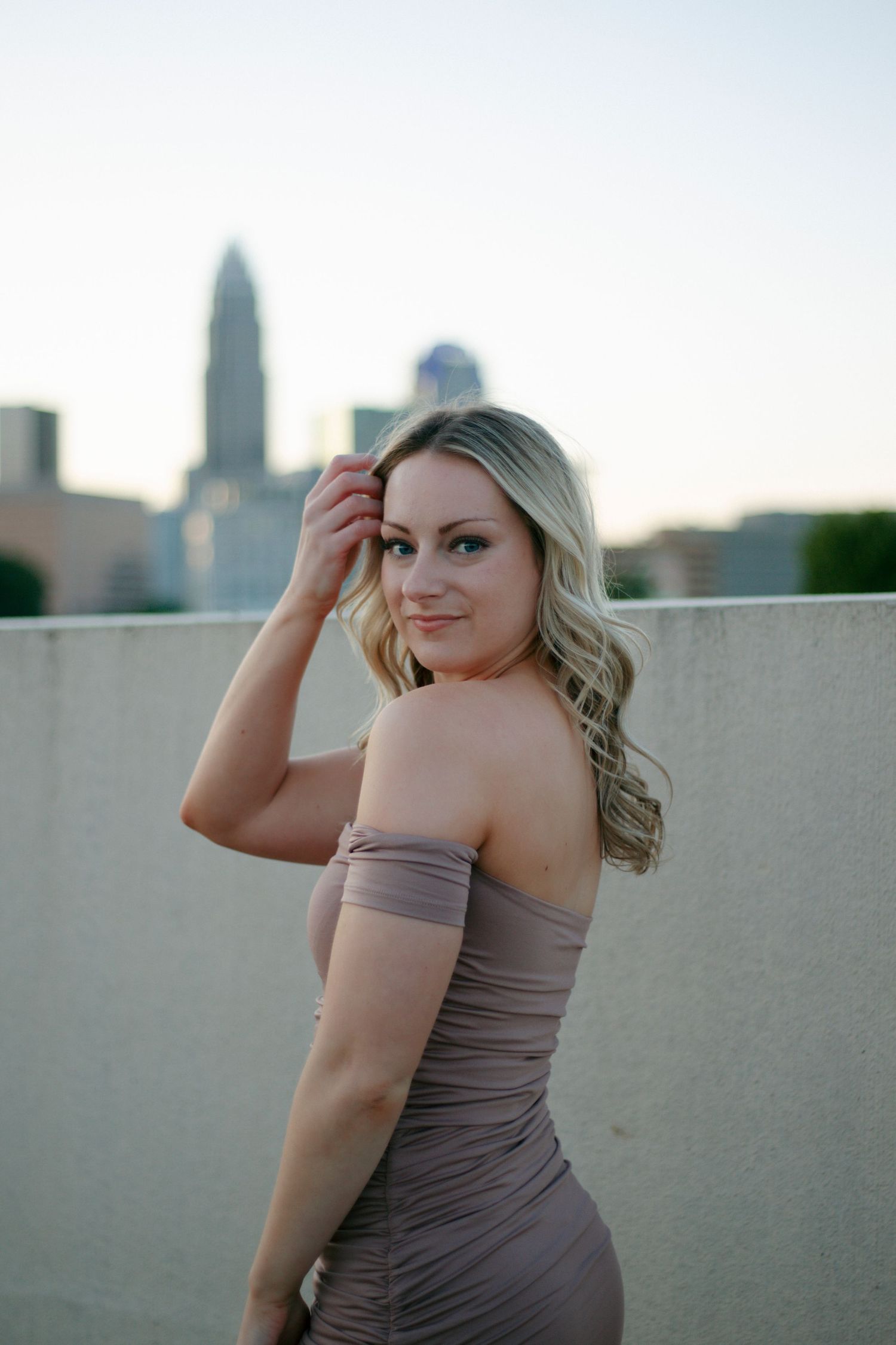 Blonde woman in off-shoulder taupe dress poses on rooftop with city skyline backdrop at dusk.