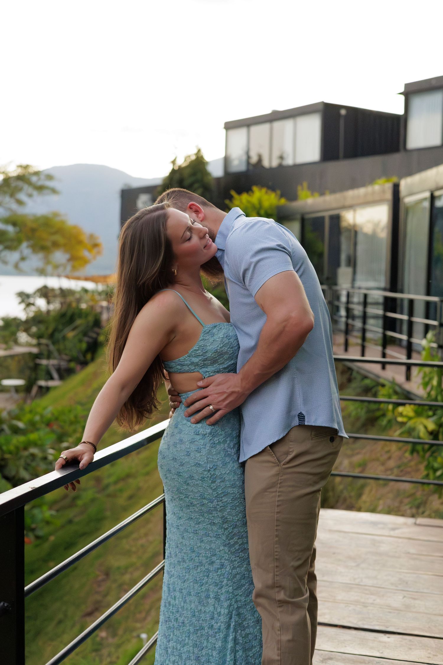 A couple kissing on a wooden deck with a modern house and scenic water view in the background.