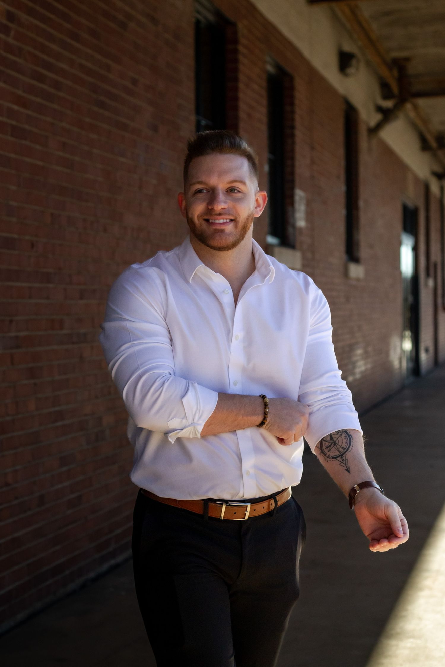Smiling man in white shirt with tattoos walks along brick building corridor in stylish business casual attire.
