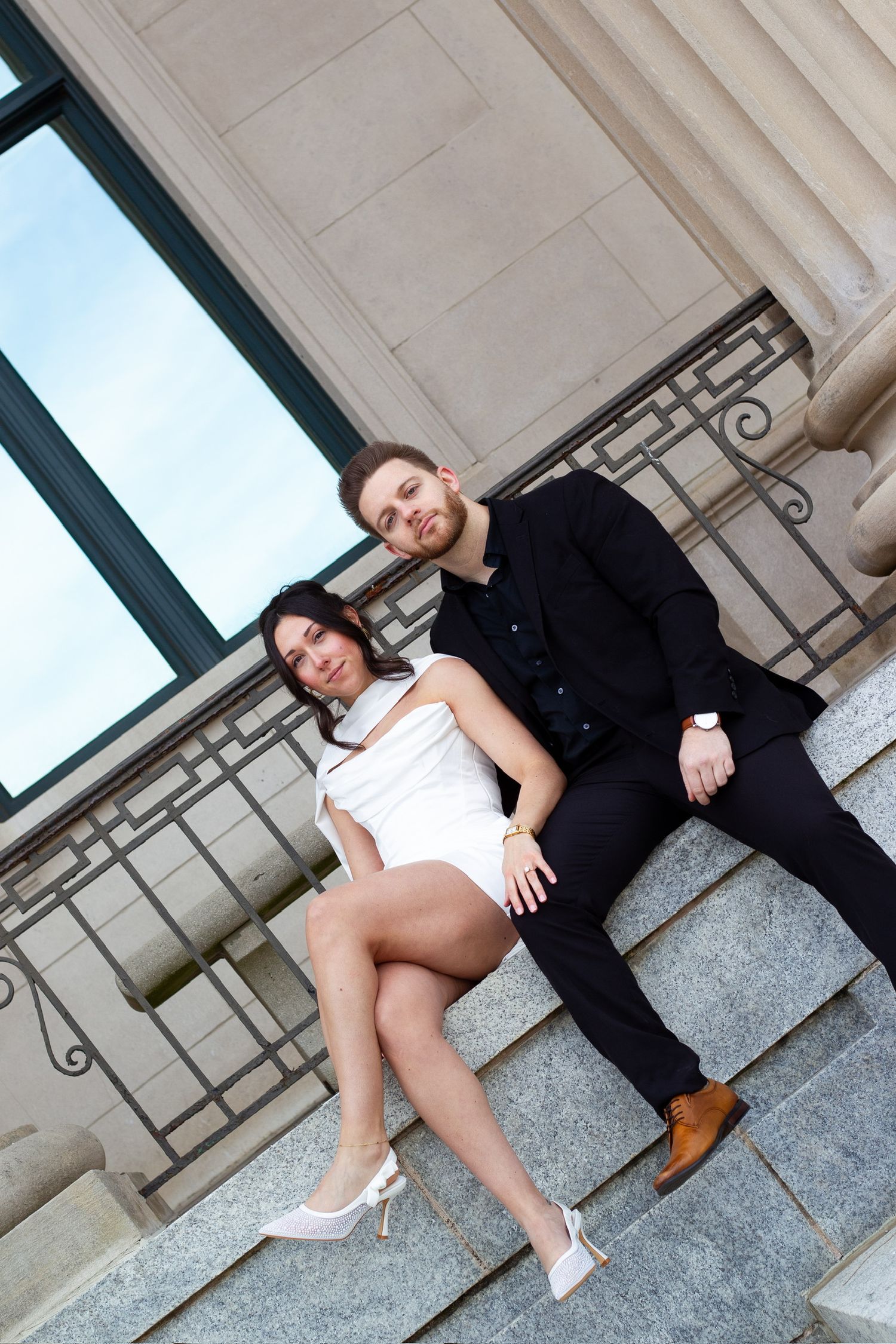 Couple posing on stone steps, woman in white dress and heels, man in black suit, elegant building backdrop.