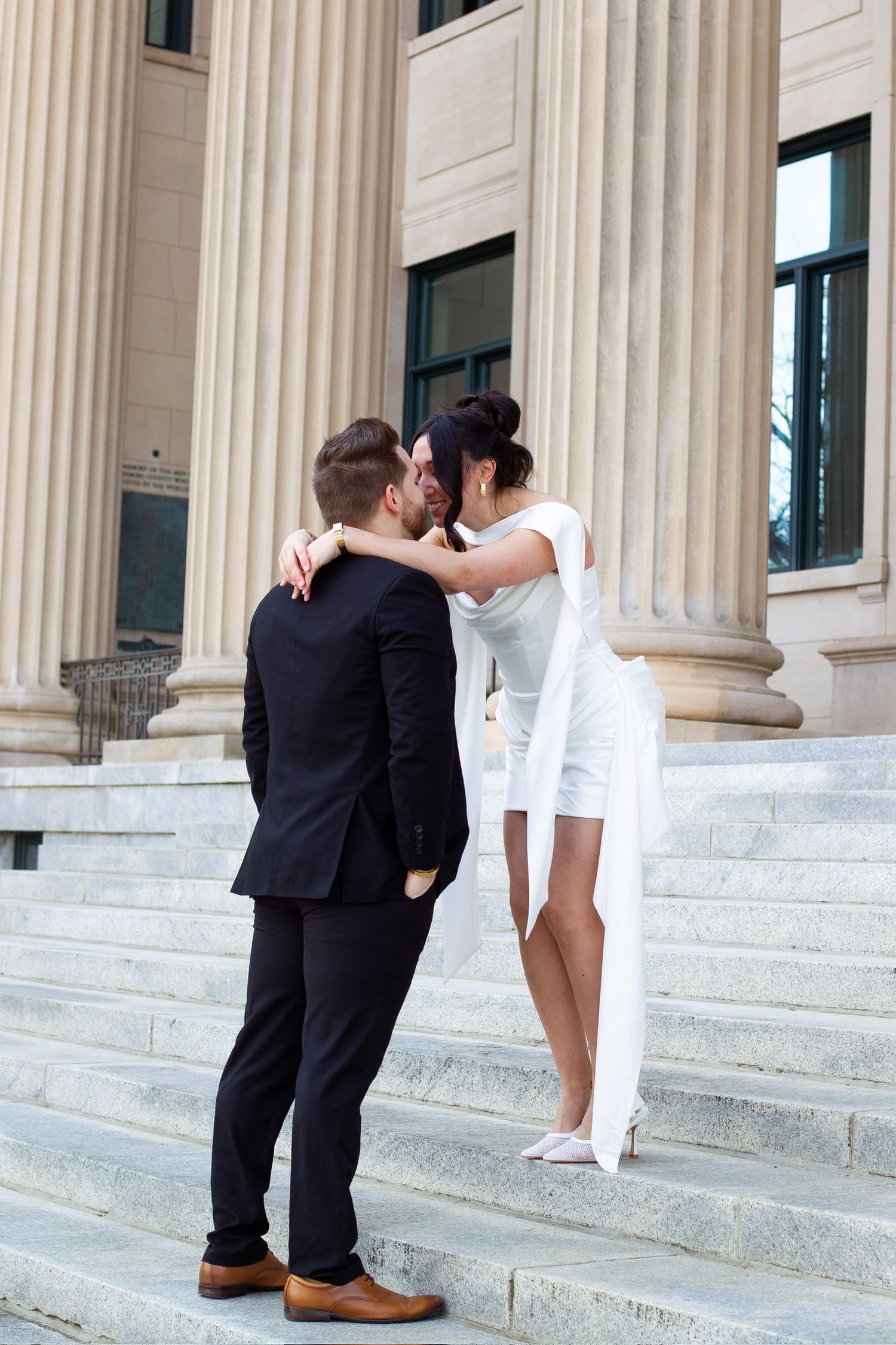 Bride in short white dress and groom in black suit embracing and kissing on steps of a columned building.