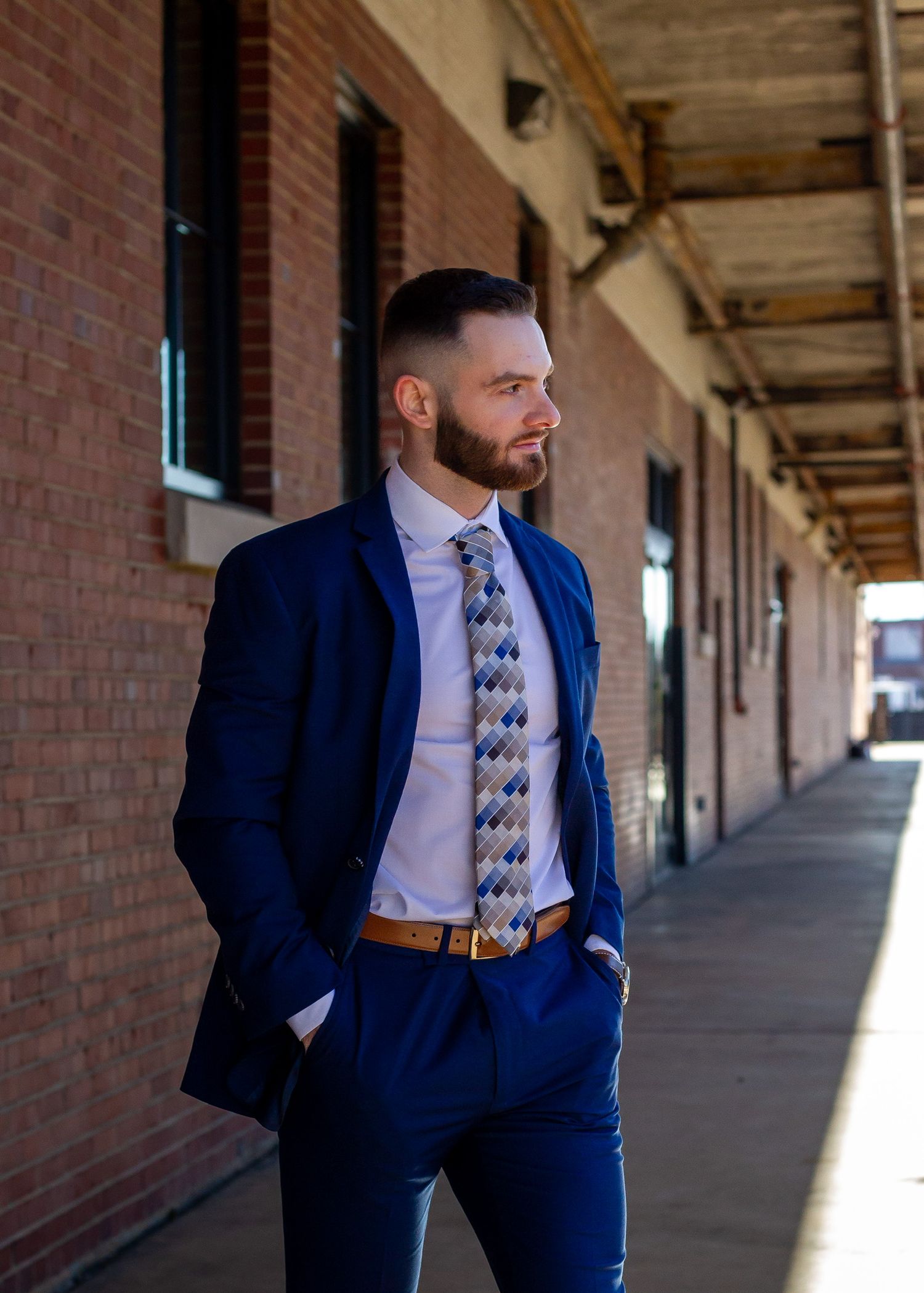 Man in navy blue suit with patterned tie and brown belt standing outside brick building.