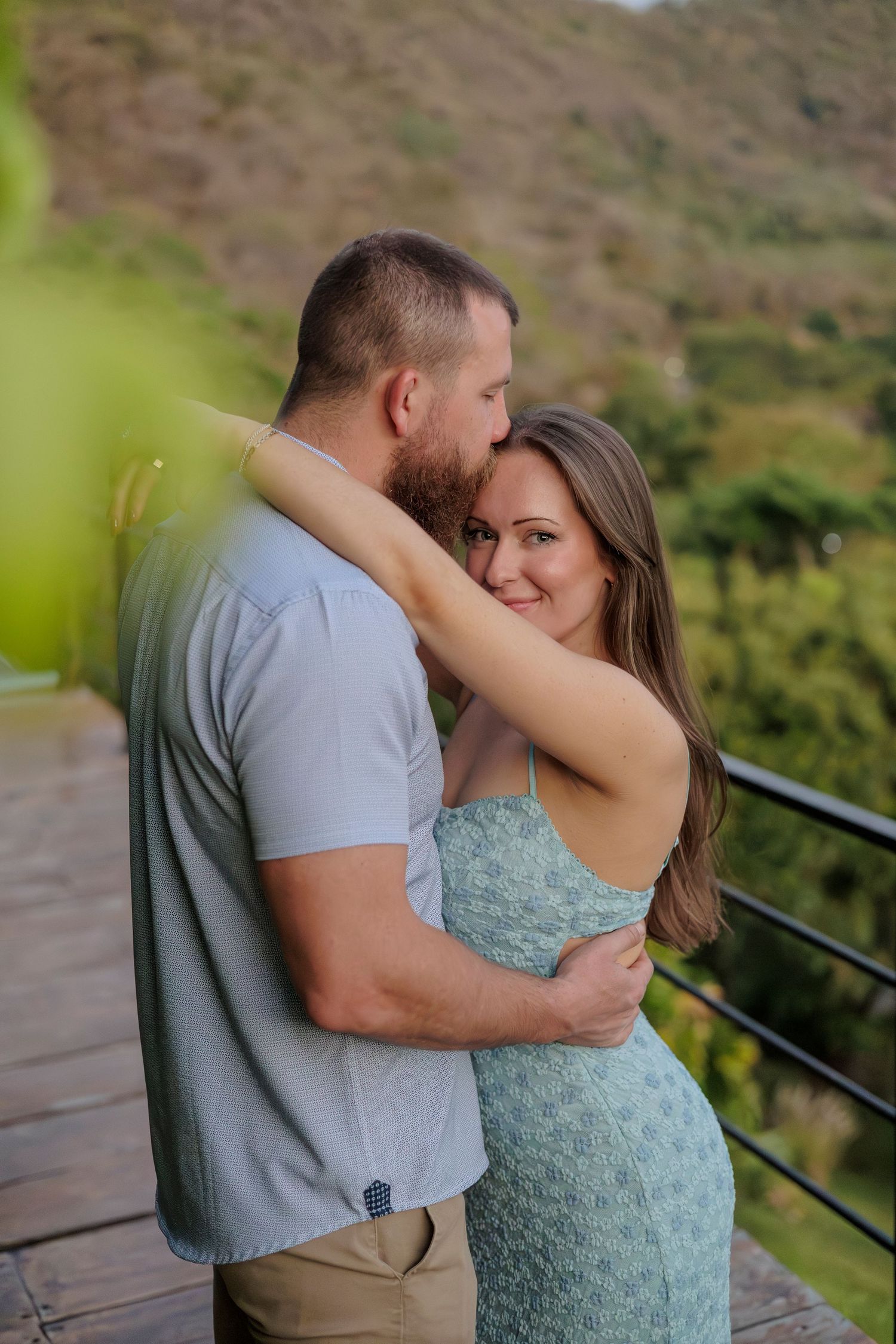 Couple embracing on a wooden deck with lush green mountains in the background, woman smiling warmly.