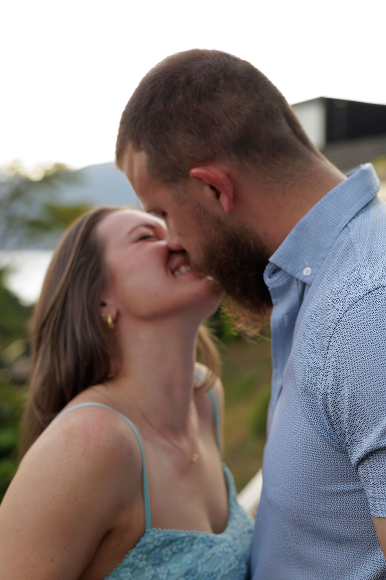 A couple sharing a romantic kiss outdoors, with the woman in a light blue dress and man in a blue shirt.