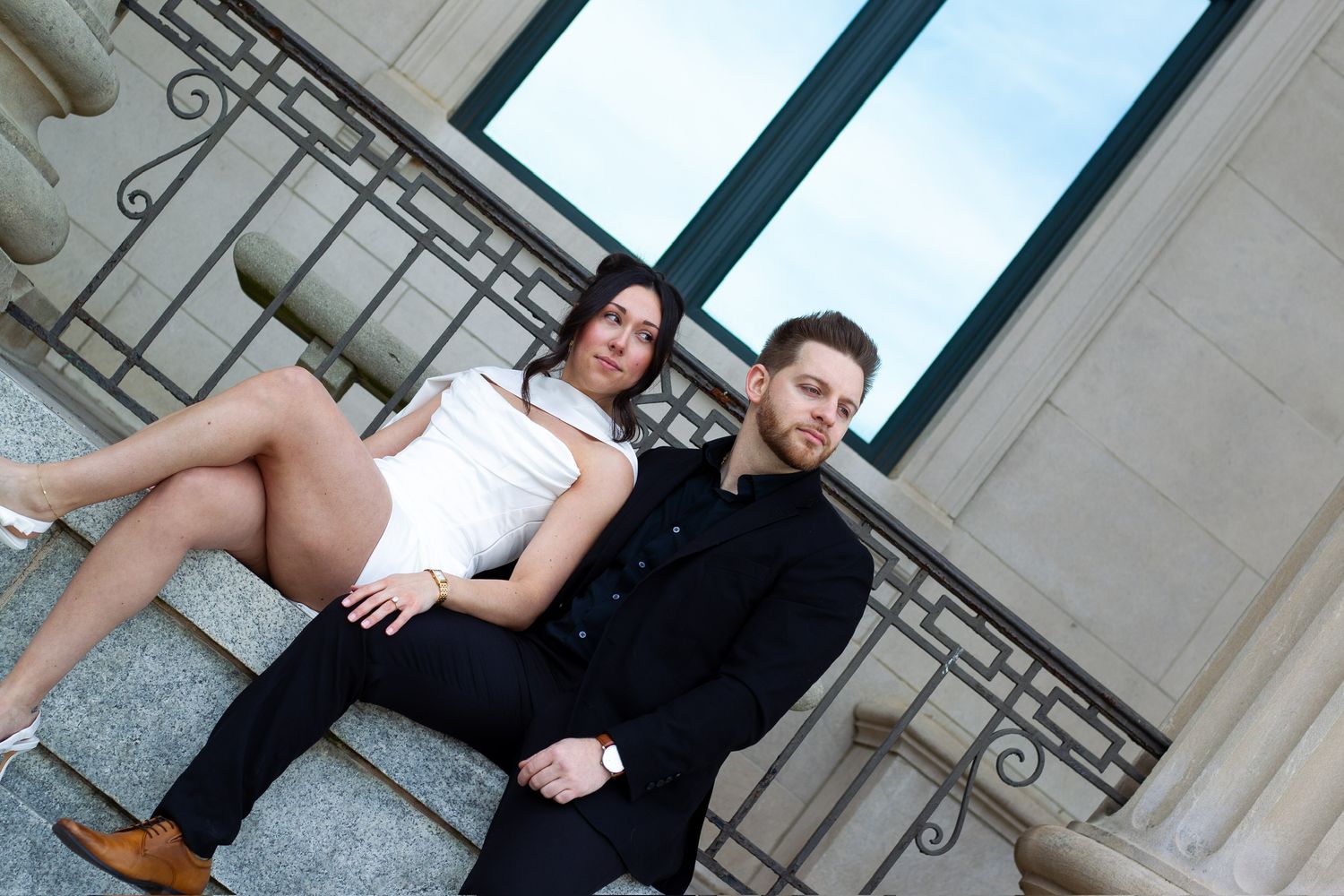 Couple posing on marble steps outdoors, woman in white top sitting on man's lap in black suit.
