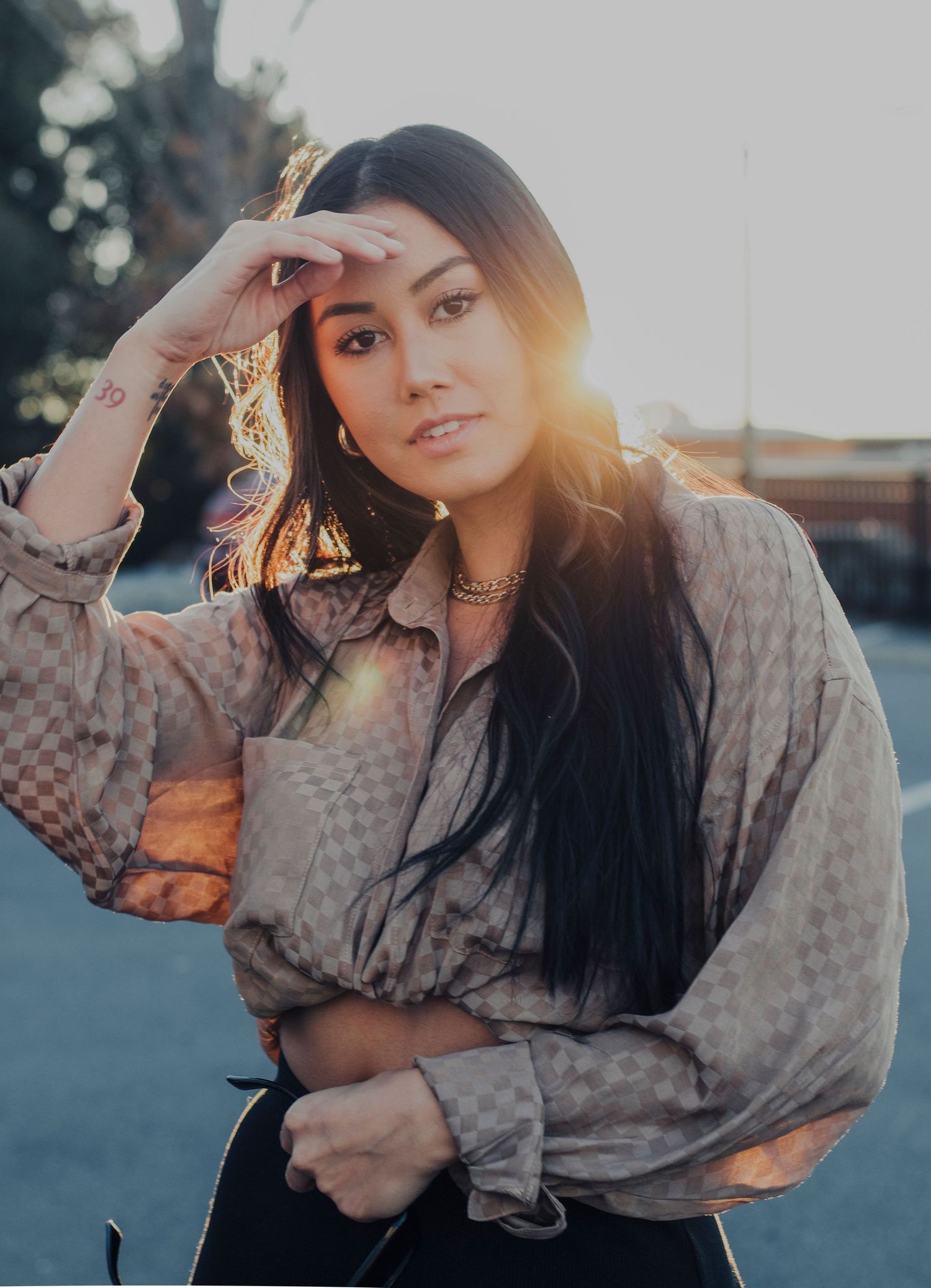 Young woman with long dark hair poses outdoors in a patterned blouse with golden sunset lens flare behind her.