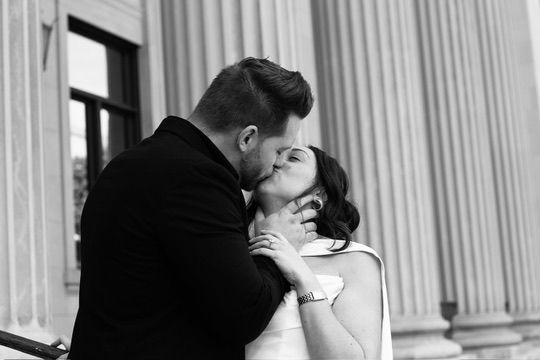 Black and white photo of a couple sharing a romantic kiss near elegant stone columns at a wedding.