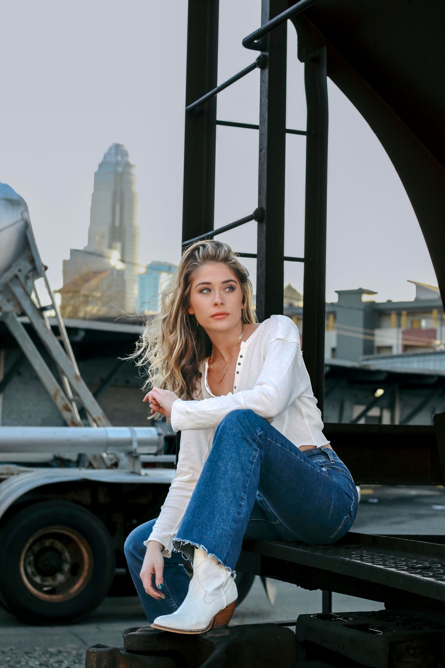 Young woman in white blouse, jeans, and cowboy boots sits on metal structure with city skyline in background.