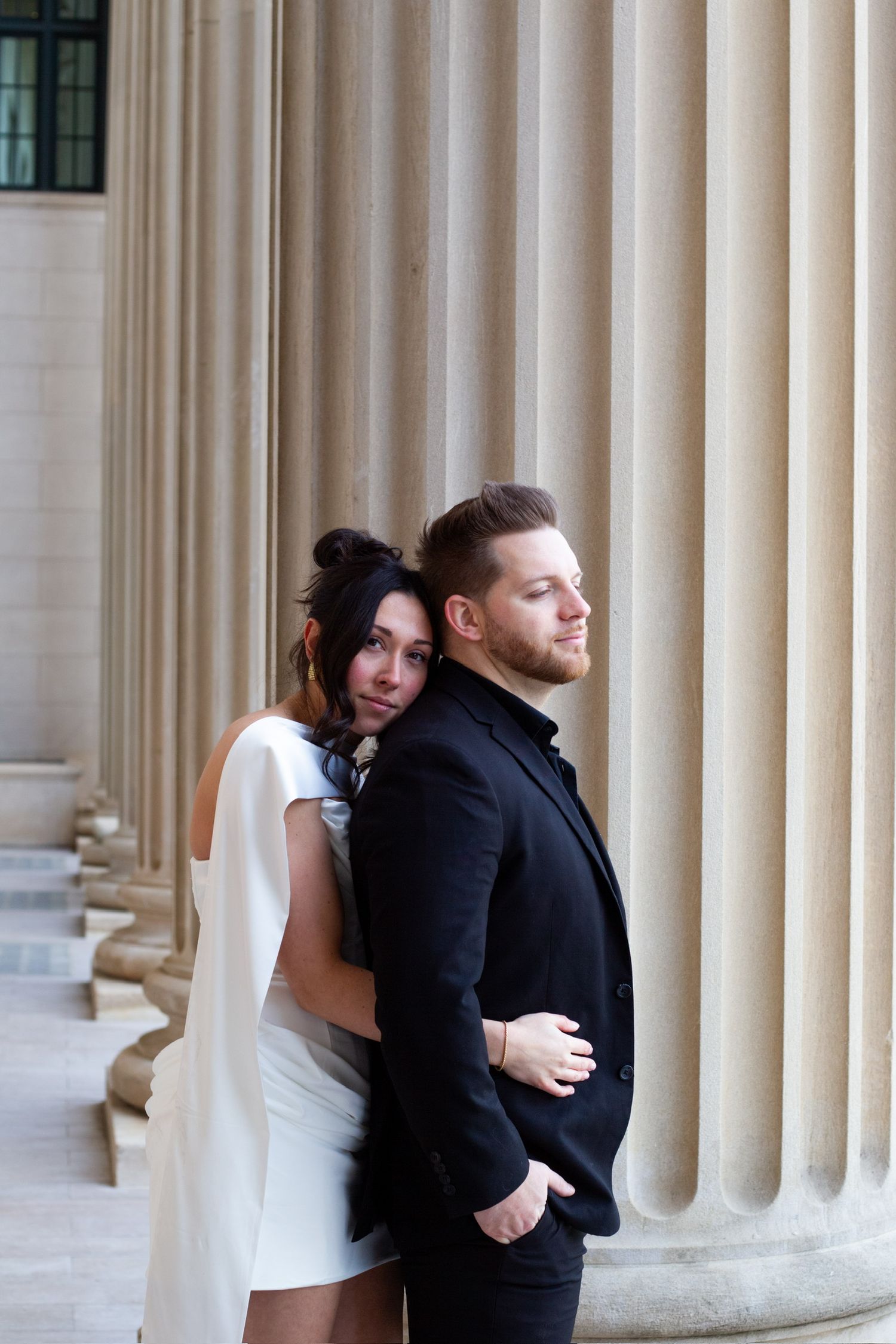A couple poses elegantly by classical columns, woman in white dress embracing man in black suit.