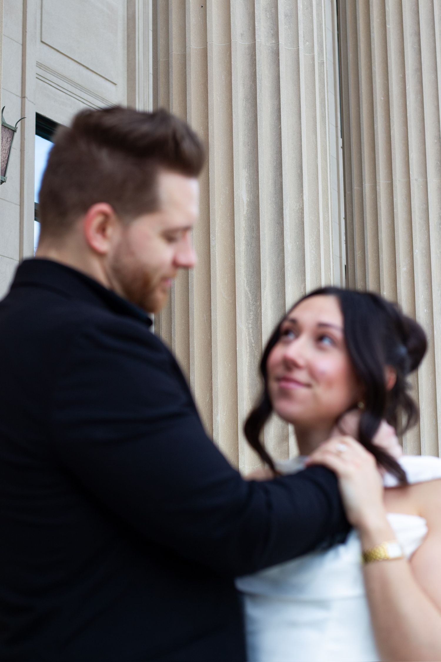 Couple sharing a romantic moment by stone columns, man in black jacket tenderly holding womans face as she gazes up smiling.