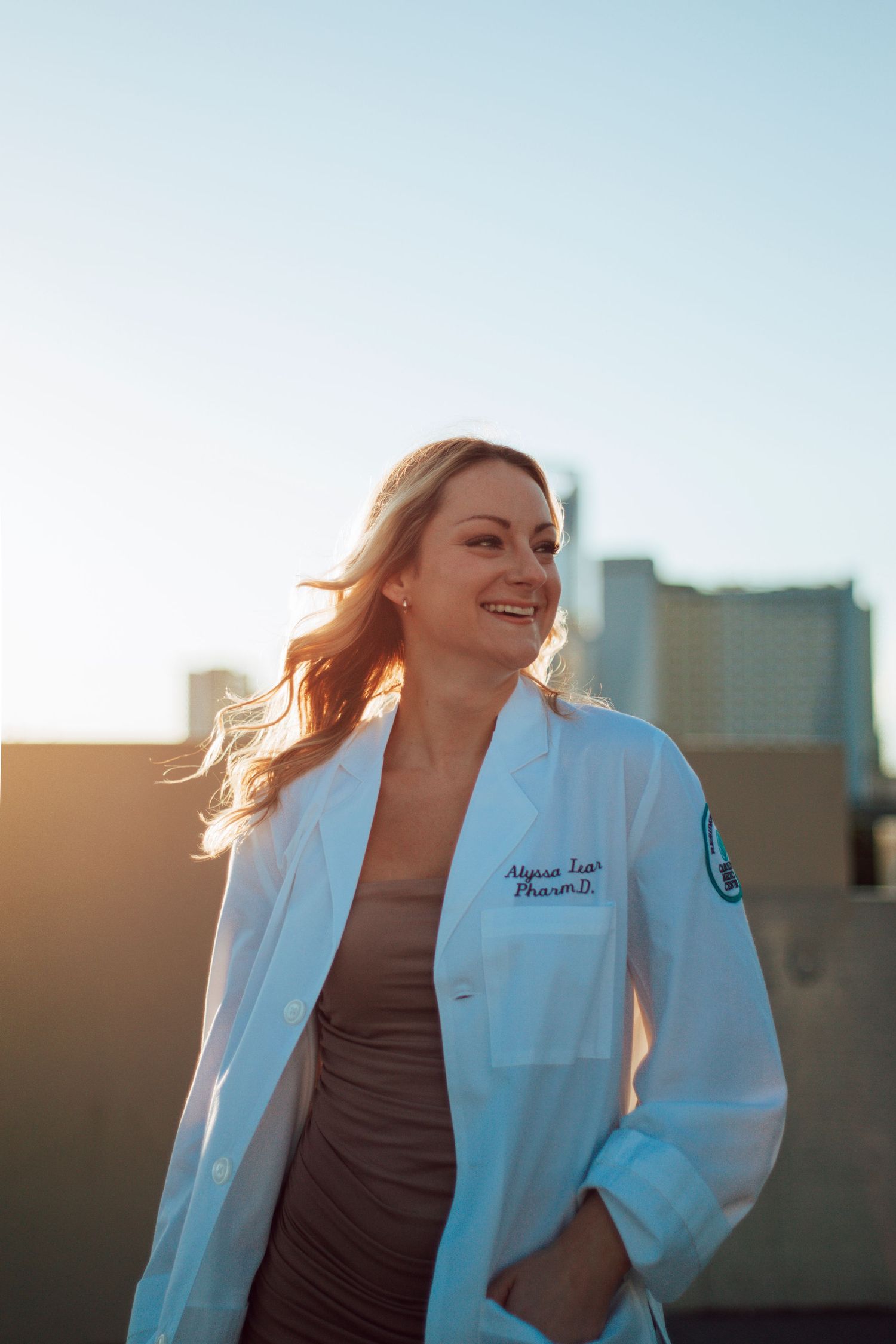 A smiling female pharmacist in a white lab coat stands outdoors at sunset with a city skyline in the background.
