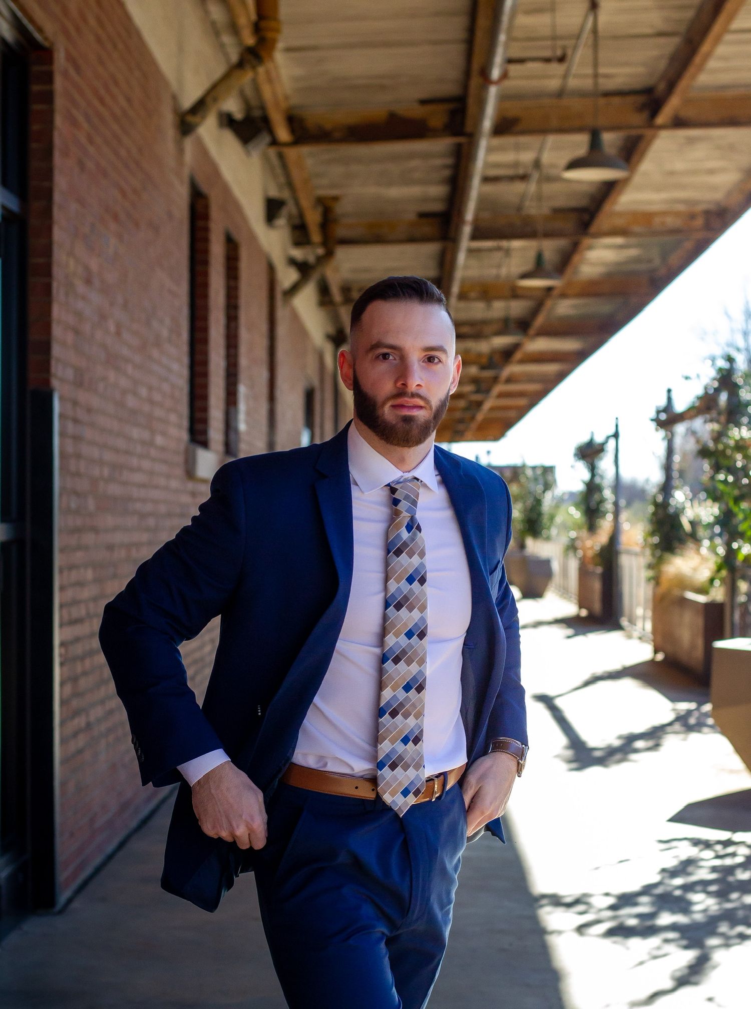 Man in navy blue suit and patterned tie walking confidently along a brick building corridor outdoors.