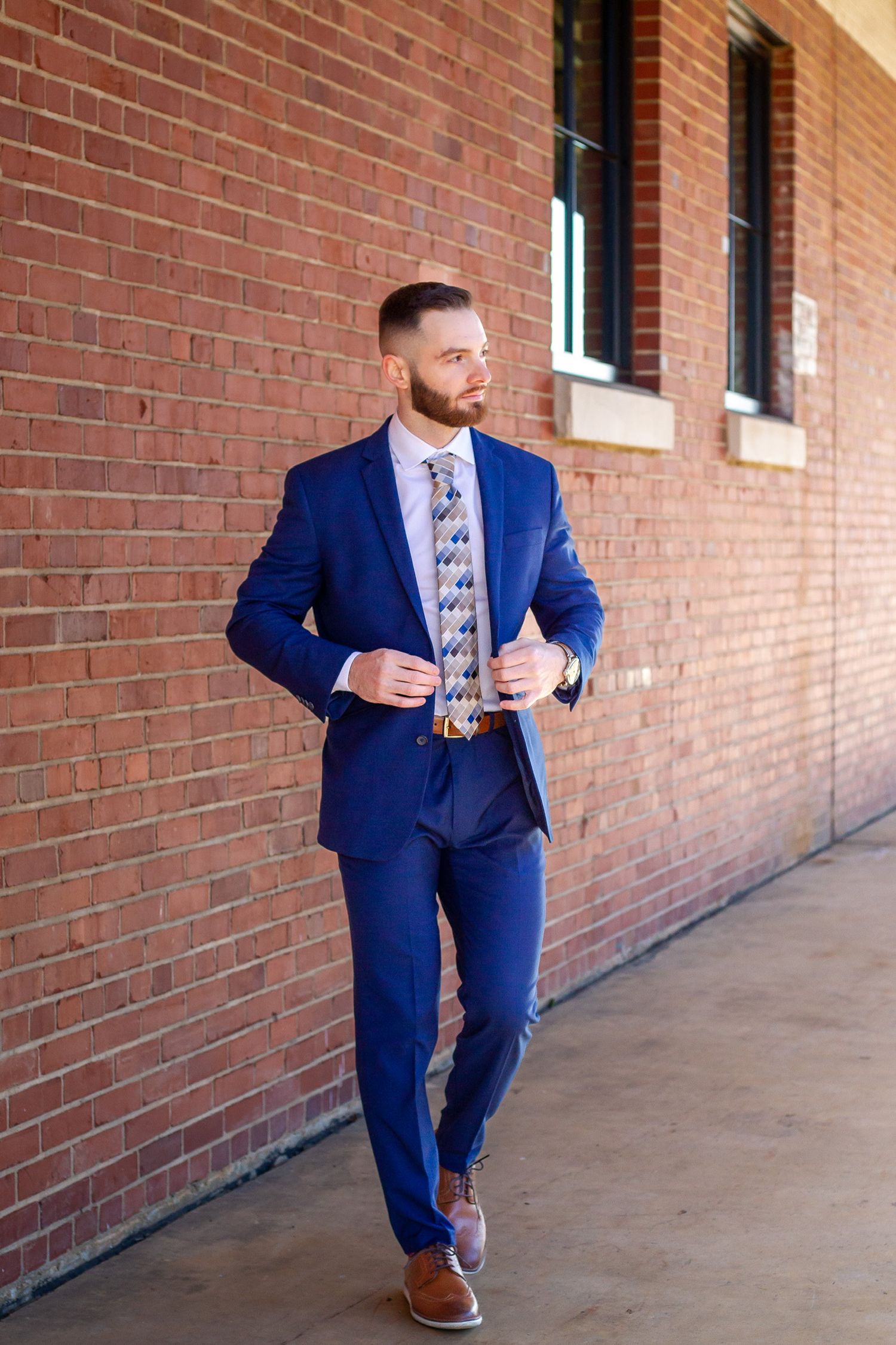 Man in blue suit with patterned tie and brown shoes walking near brick wall.