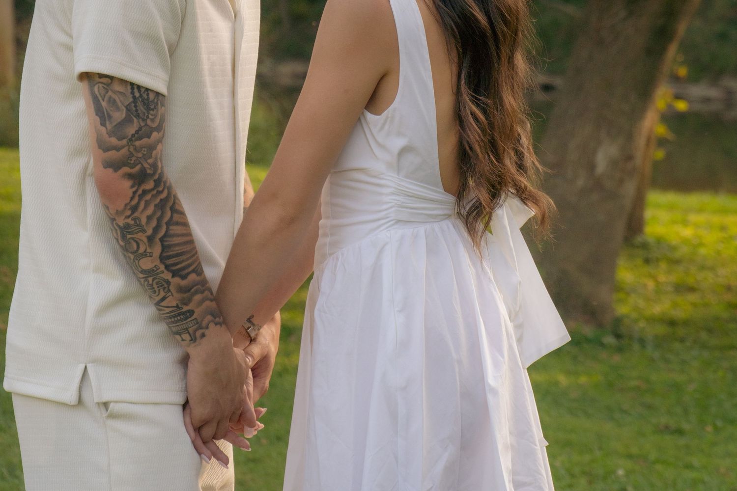 A couple in white outfits embraces tenderly in an outdoor setting with trees and golden sunlight.