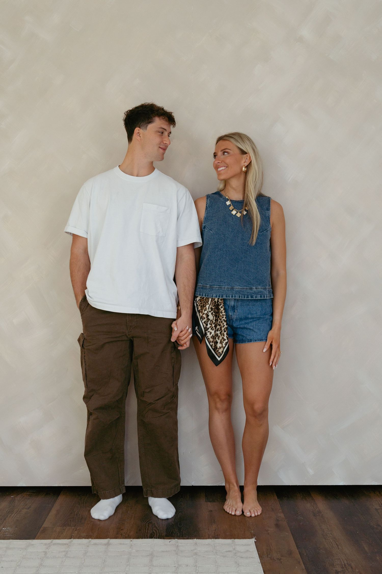 Couple in casual wear poses together against white wall in minimalist style photoshoot.