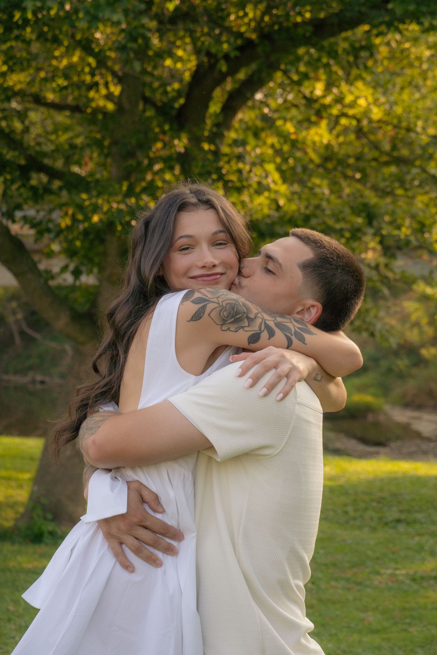 A romantic embrace captured in a sunny park with lush green trees in the background.