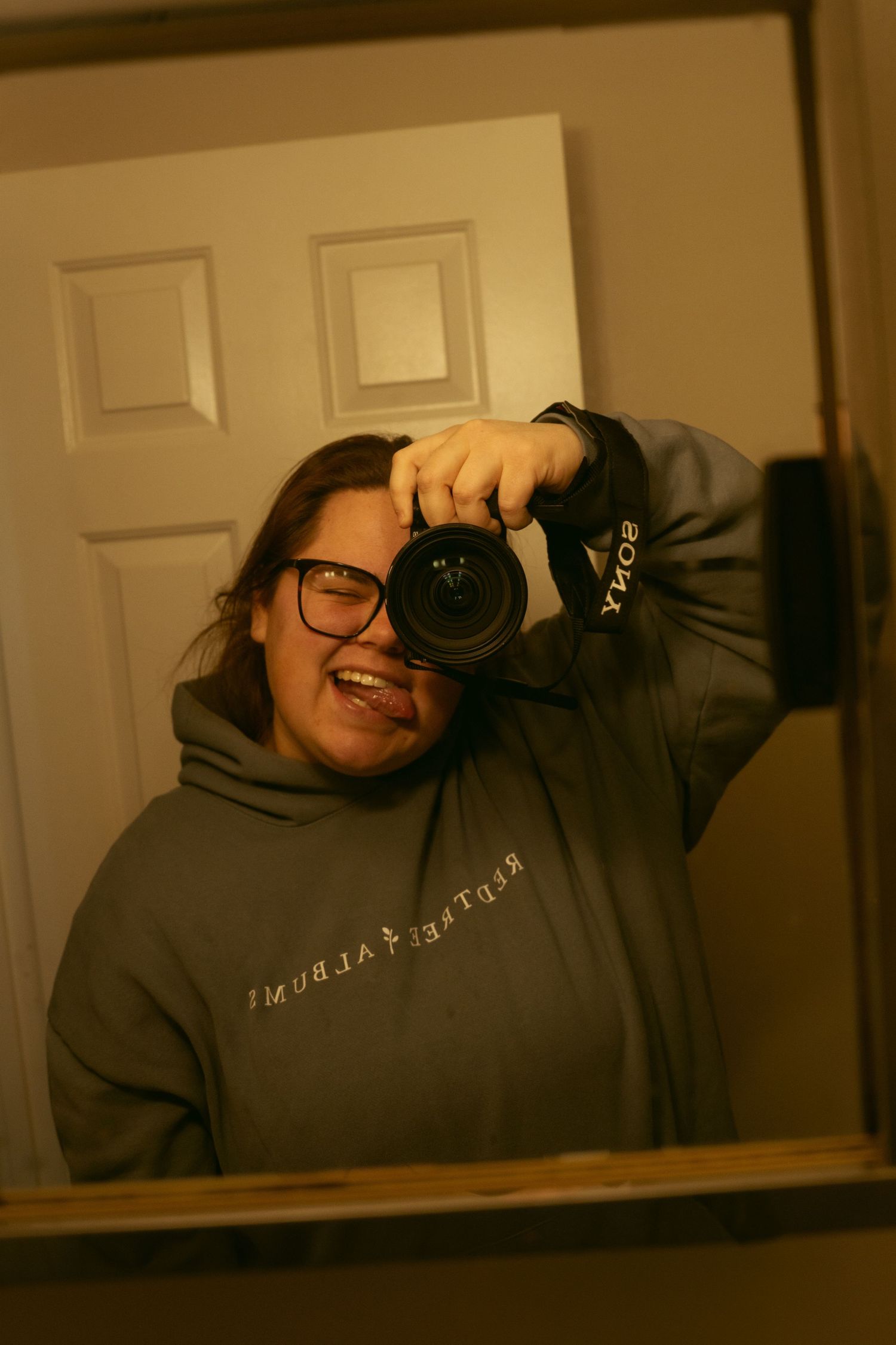Someone takes selfies with a camera while wearing a gray hoodie in dim lighting.