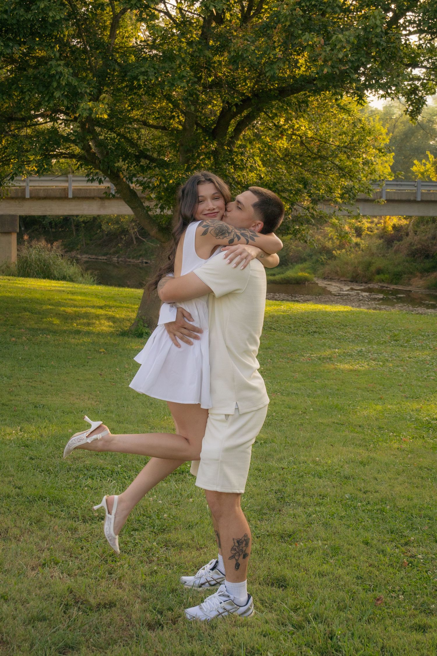 A playful couple spins and embraces on grass during golden hour in a park setting.