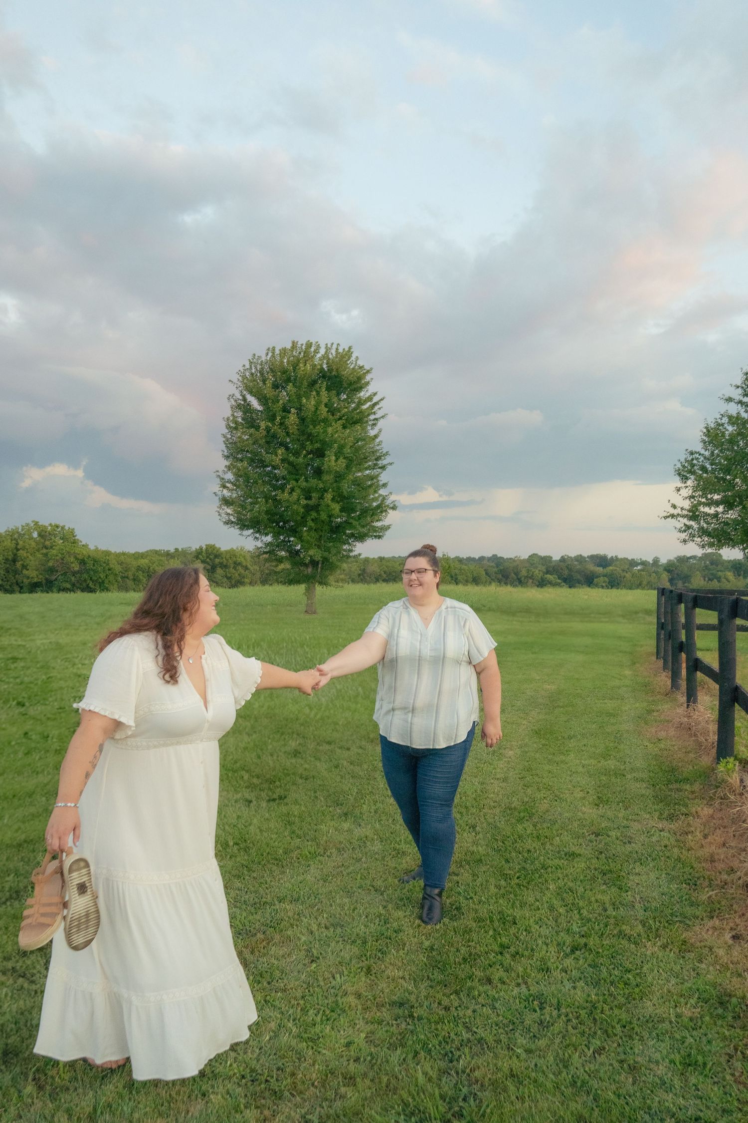Couple in white dresses holding hands while walking through a sunny meadow.