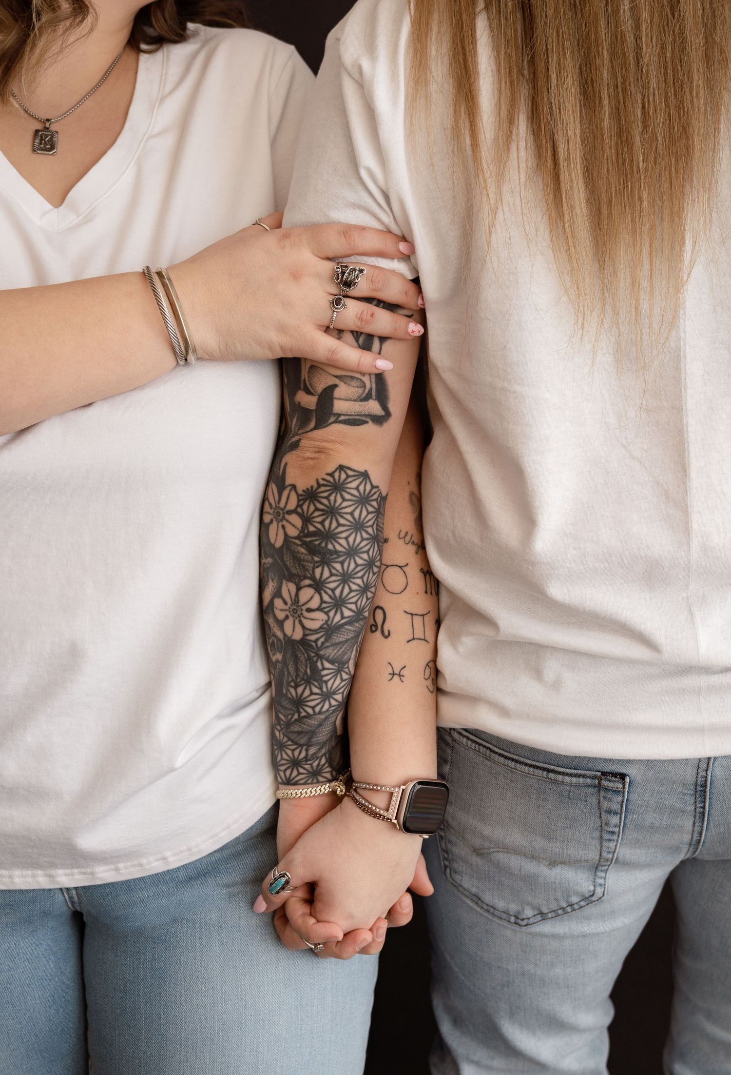 Close-up of tattooed arms and hands holding each other while wearing white shirts and jeans.