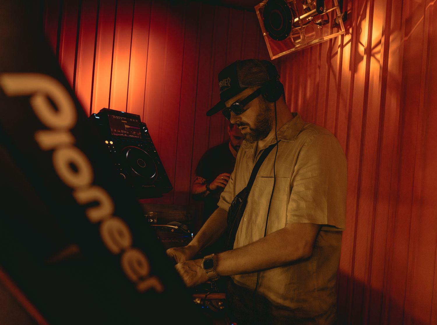 DJ working Pioneer decks in dimly lit red-toned venue wearing hat.