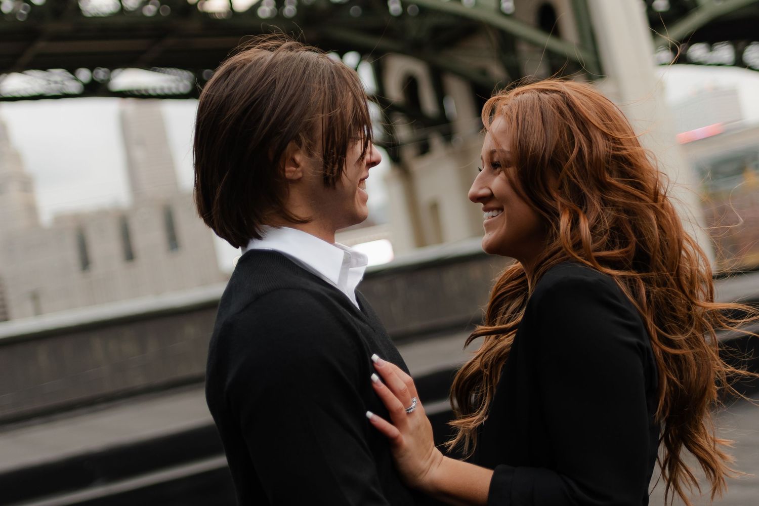 A couple shares a romantic moment under a bridge, the woman smiling with flowing red hair, both dressed in black.