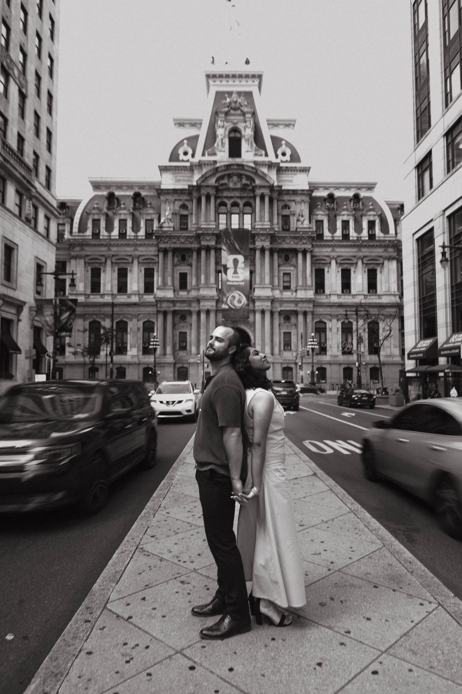 Black and white photo of a couple standing on a city sidewalk in front of Philadelphia City Hall with traffic passing by.