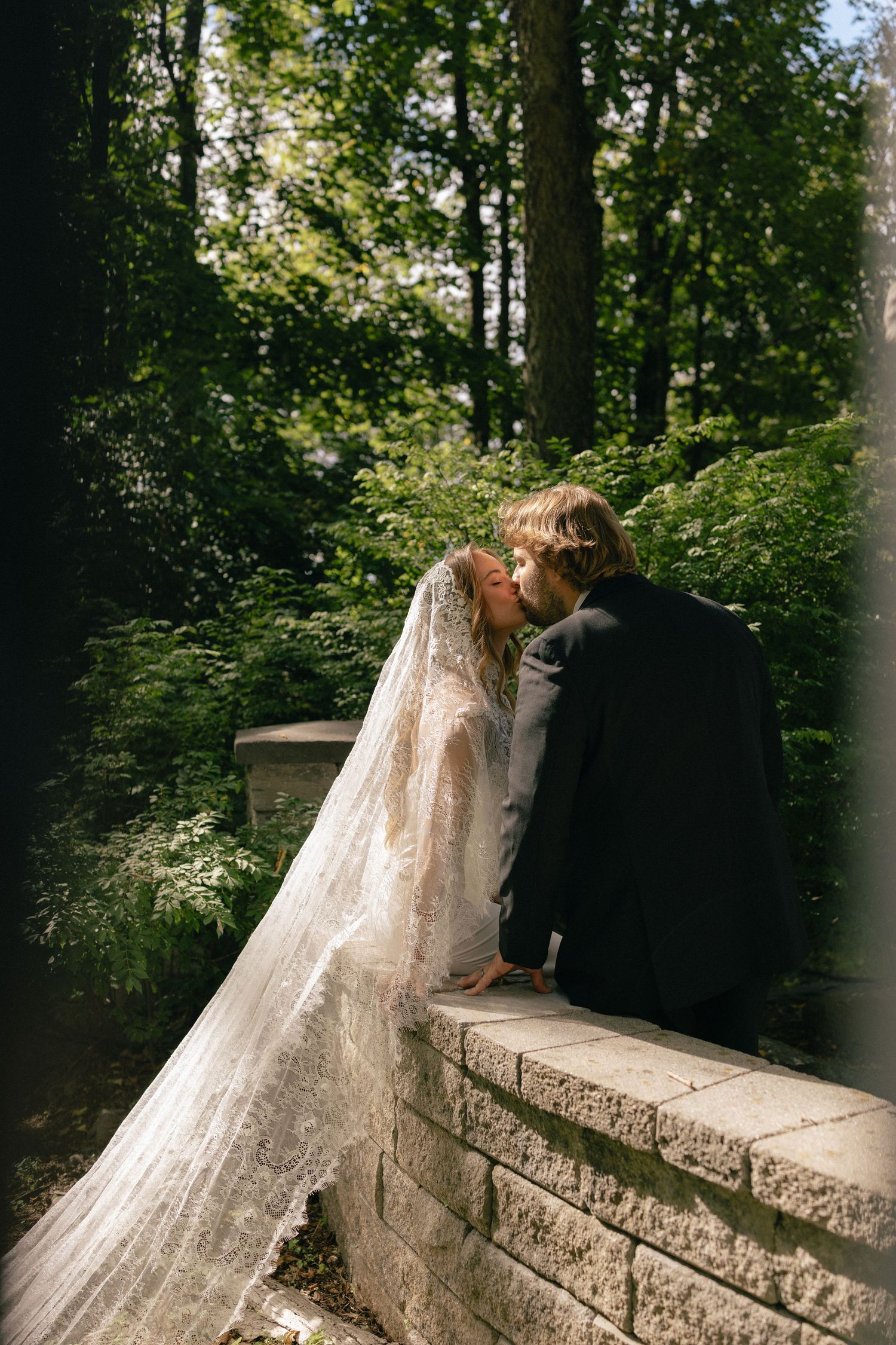 Bride and groom share a romantic kiss leaning on a stone wall surrounded by lush green trees in an outdoor wedding setting.
