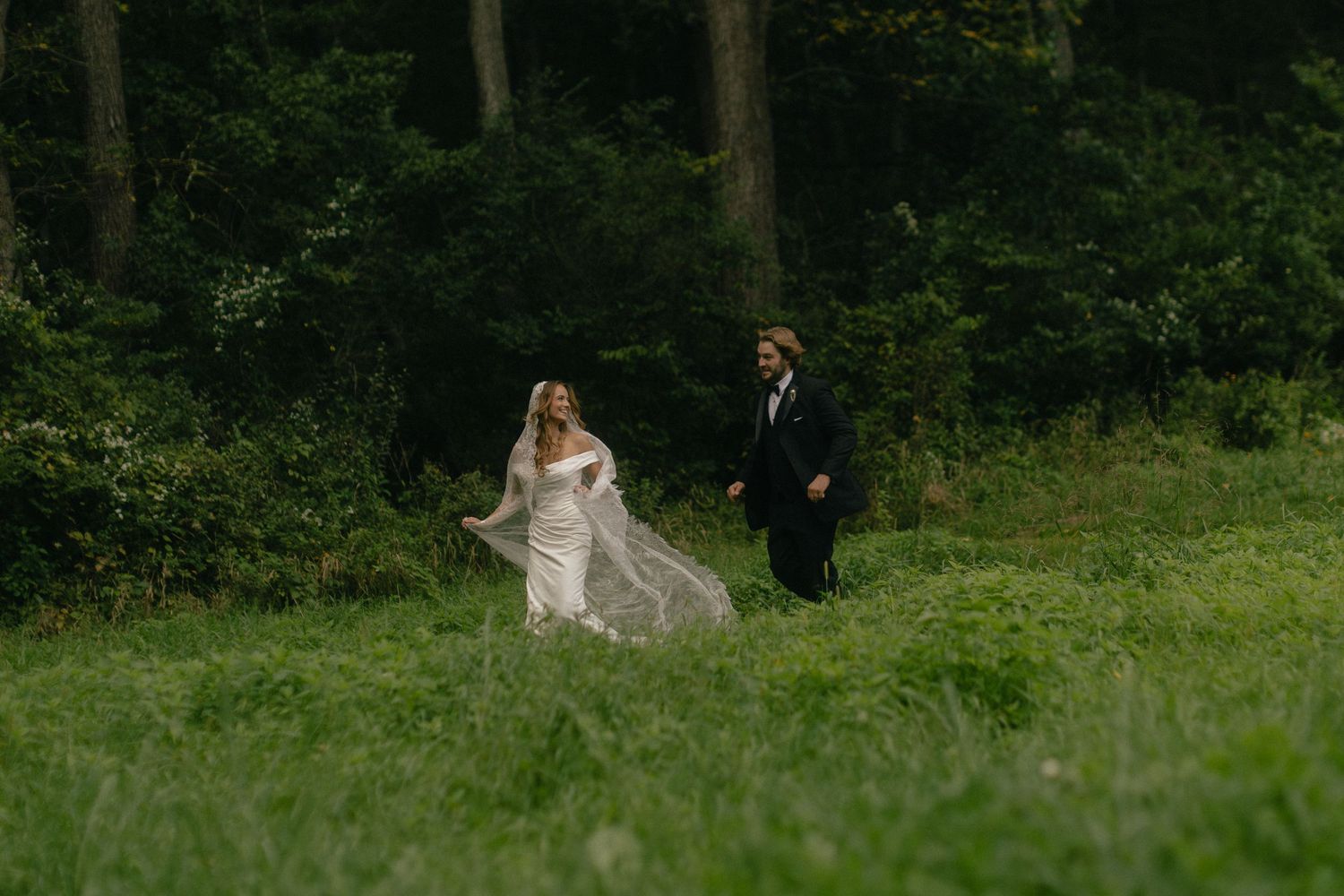 Bride in flowing white gown and groom in black suit walking through lush green meadow with forest backdrop.