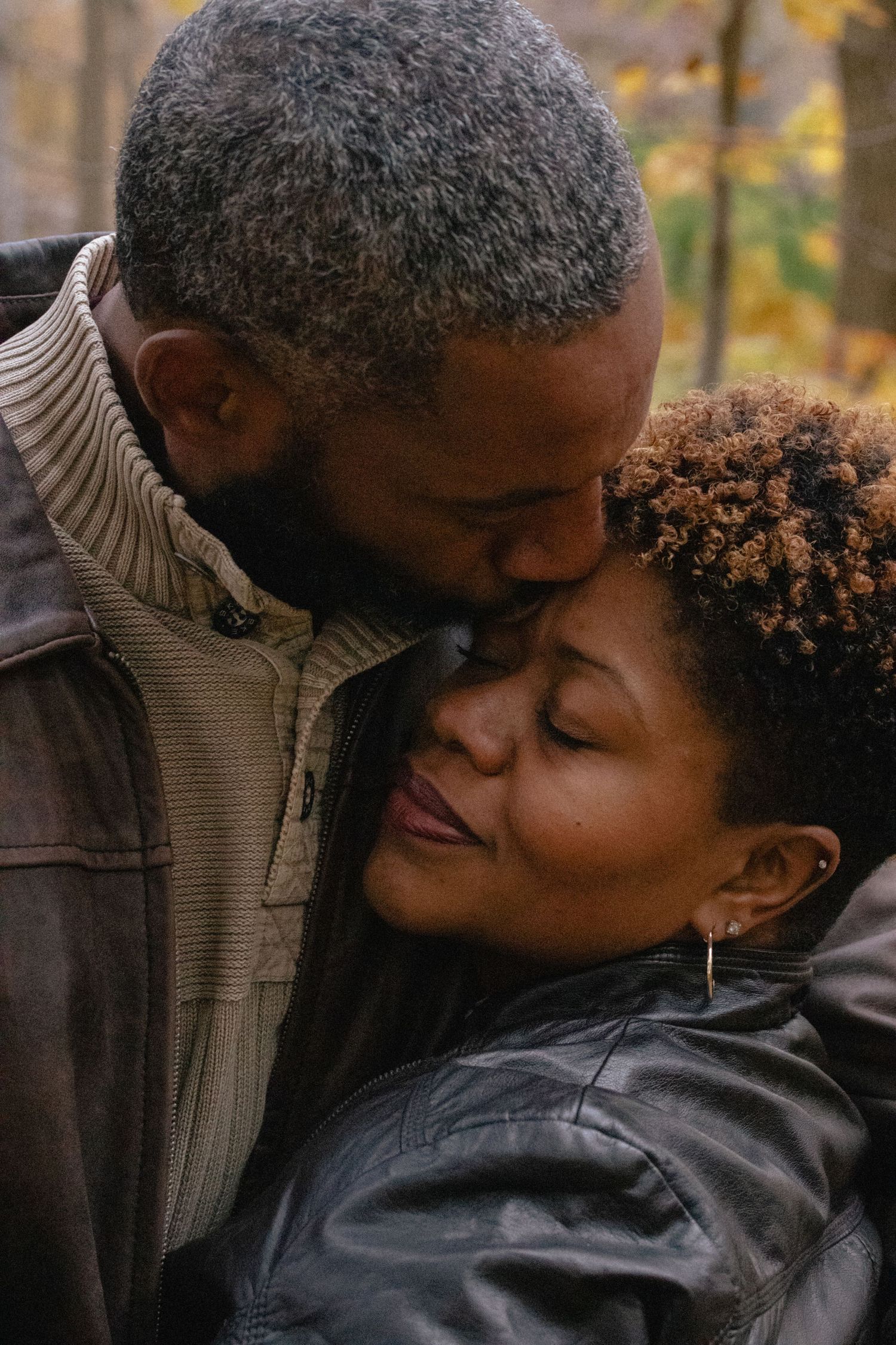 A loving Black couple shares a tender kiss outdoors in autumn, the man gently kissing the smiling woman's forehead.