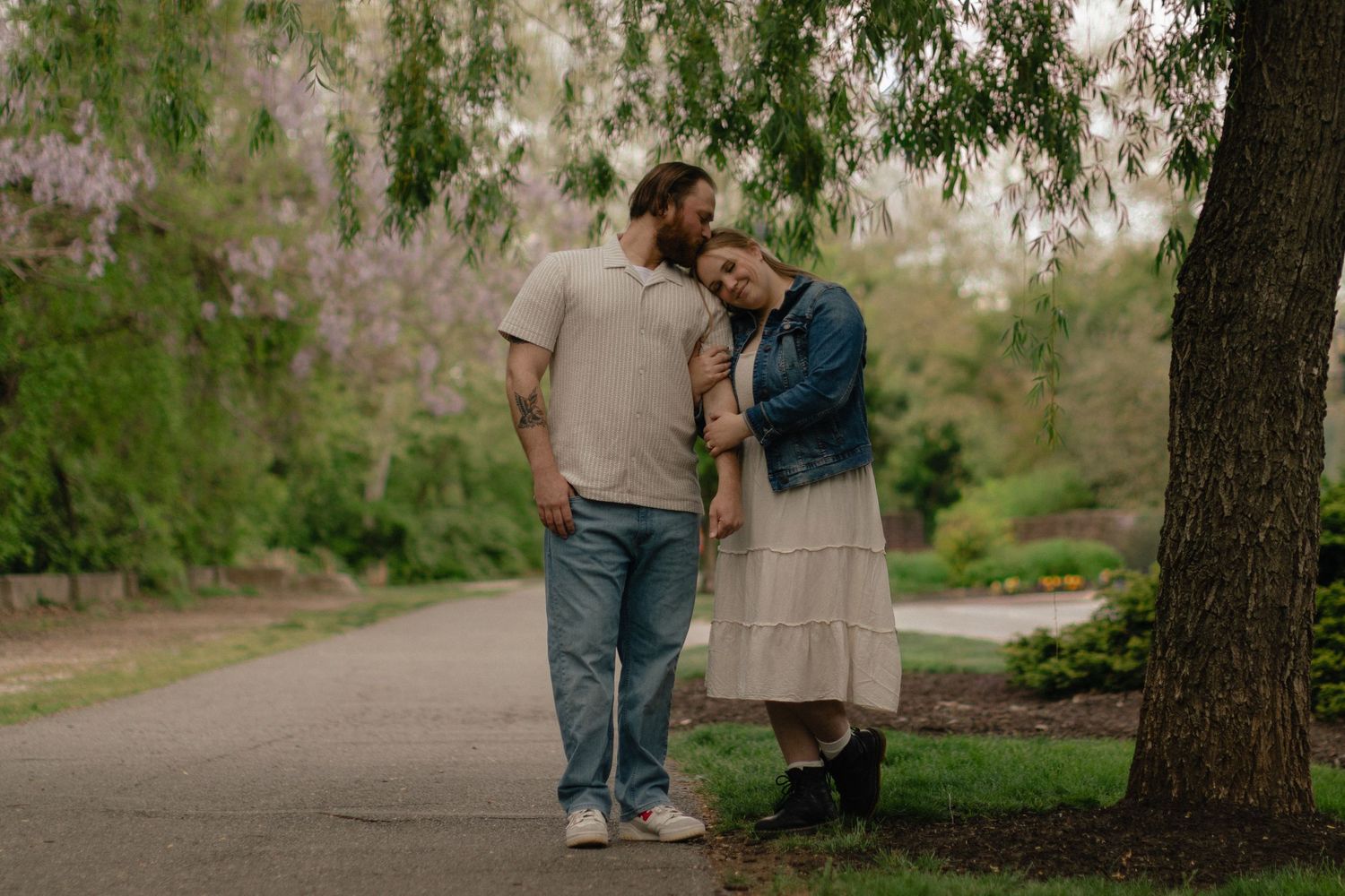 Couple embracing under a blooming tree on a park path in spring, wearing casual denim and cream outfits.