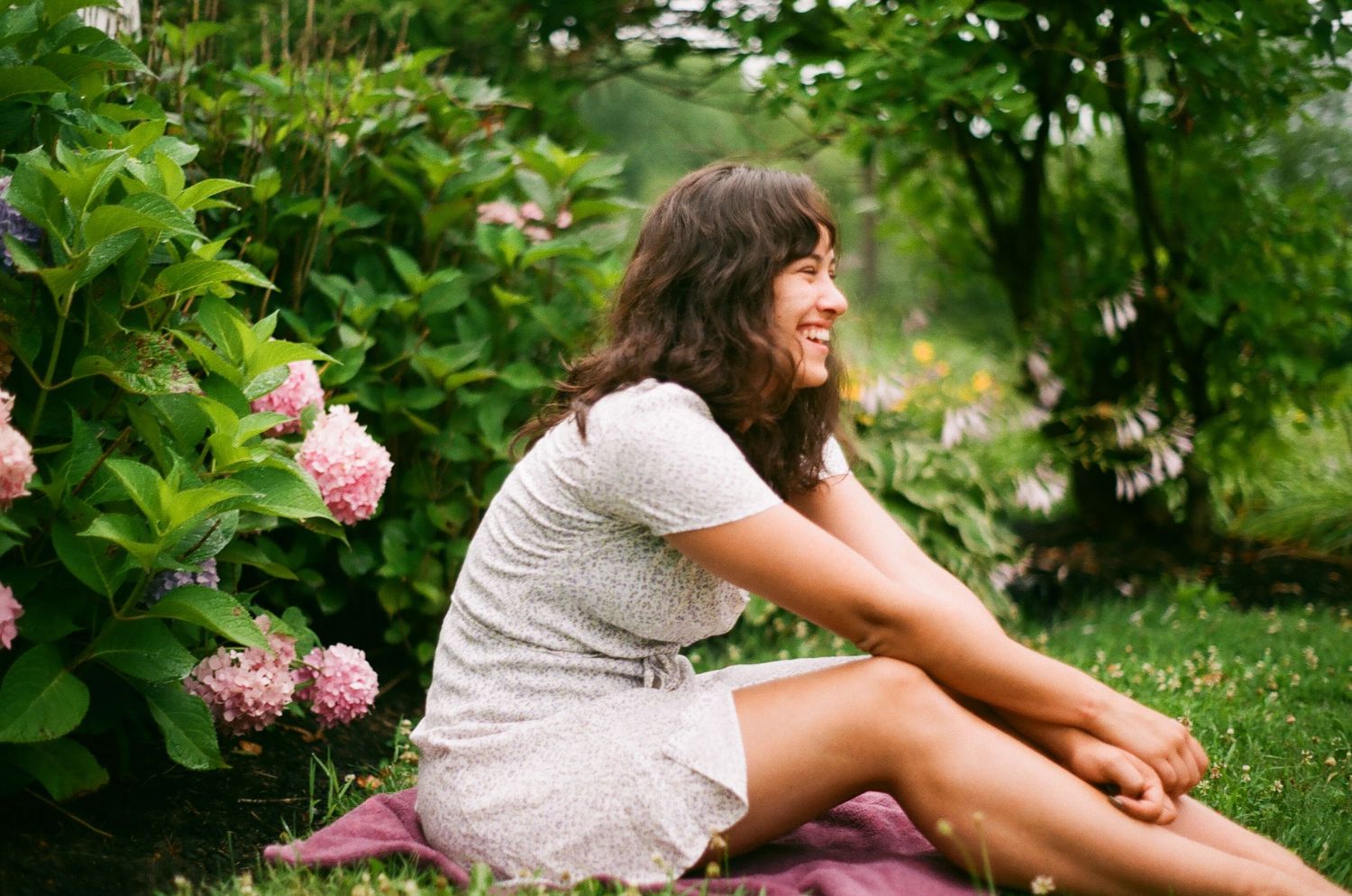 A young woman in a floral dress sits smiling in a lush garden with pink hydrangeas on a sunny day.