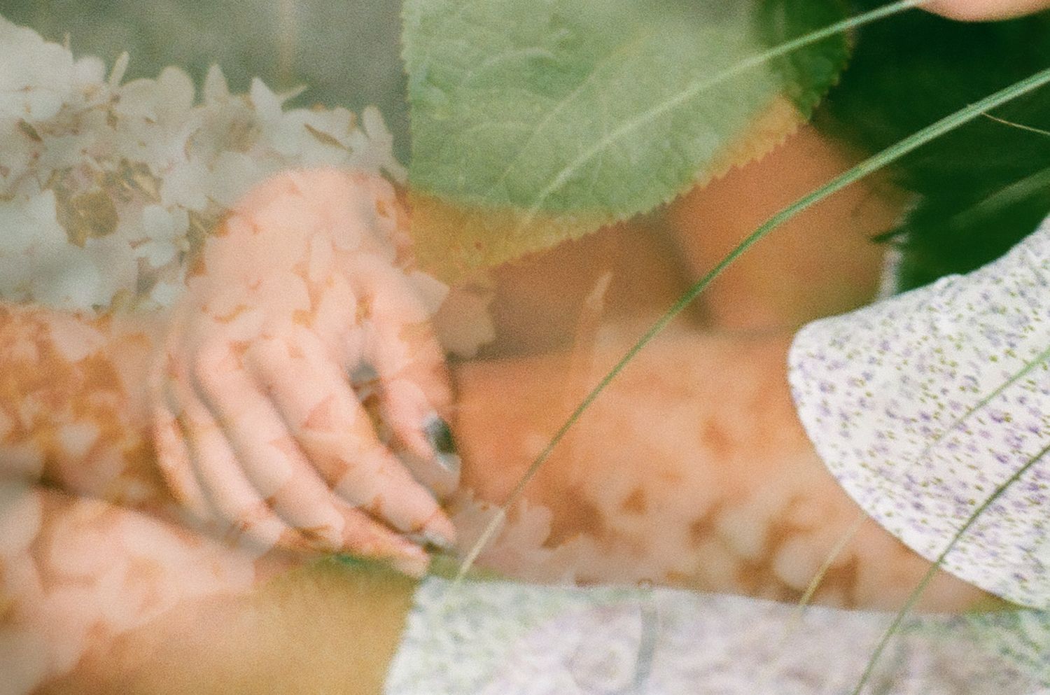 Double exposure of hands with dark nail polish overlaid with green leaves and soft floral fabric.