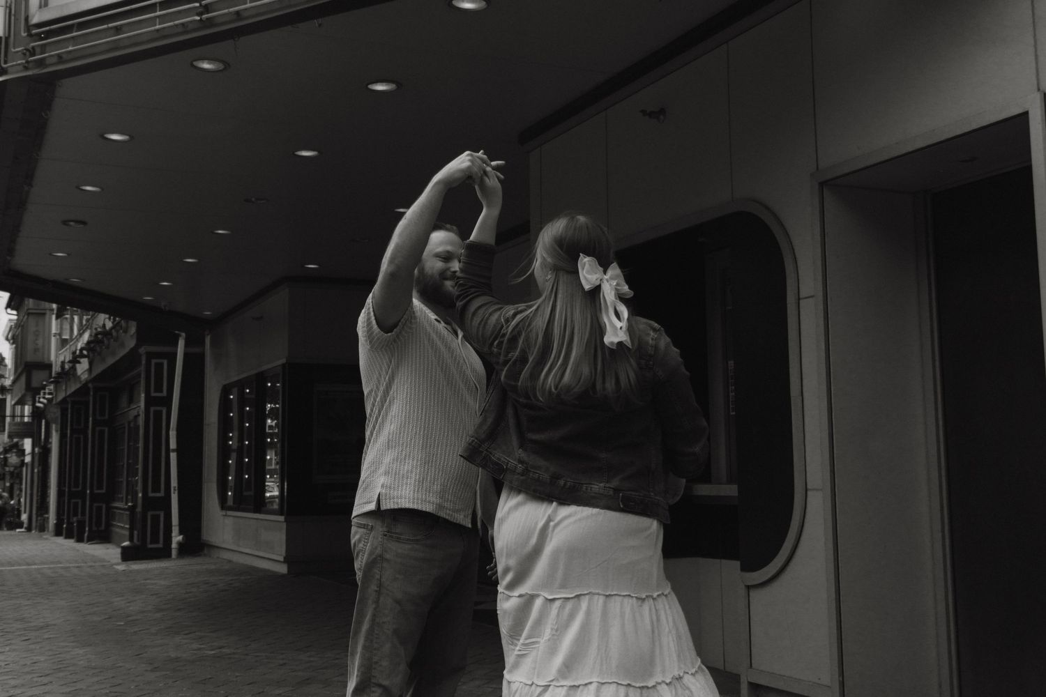 A couple dances together on a city sidewalk in black and white, the woman wearing a white skirt.